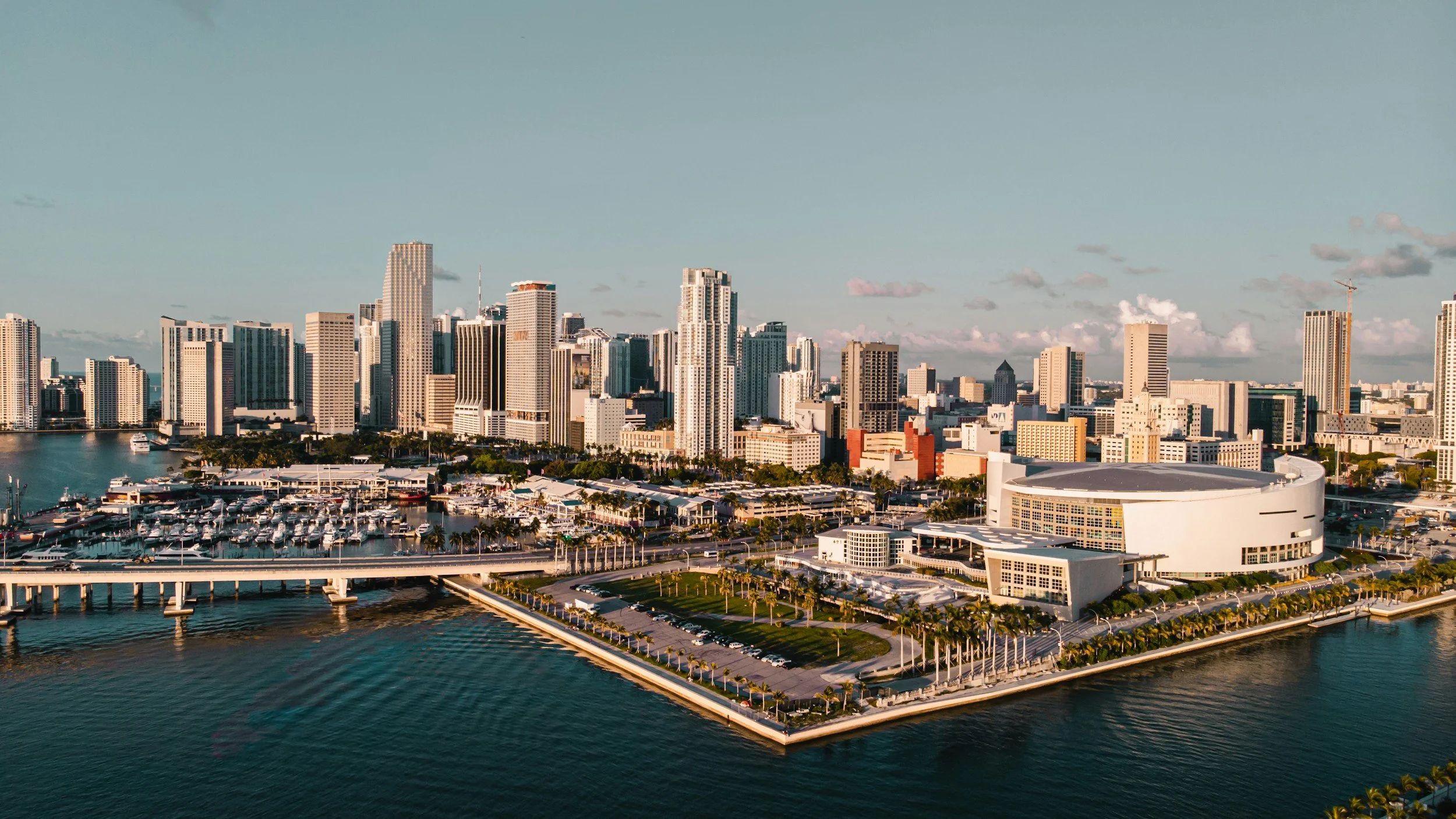 Aerial view of downtown Jacksonville, Florida, featuring tall modern skyscrapers, a river with a bridge, and a clear sky at sunset.