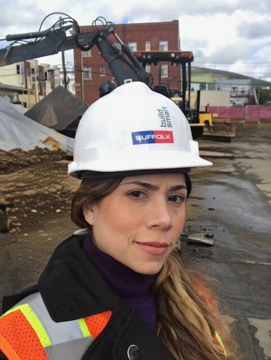 A woman in construction gear, wearing a white hard hat with a 'SUFFOLK build SMART' sticker, and a high-visibility safety vest, stands at a construction site with heavy machinery and buildings in the background.
