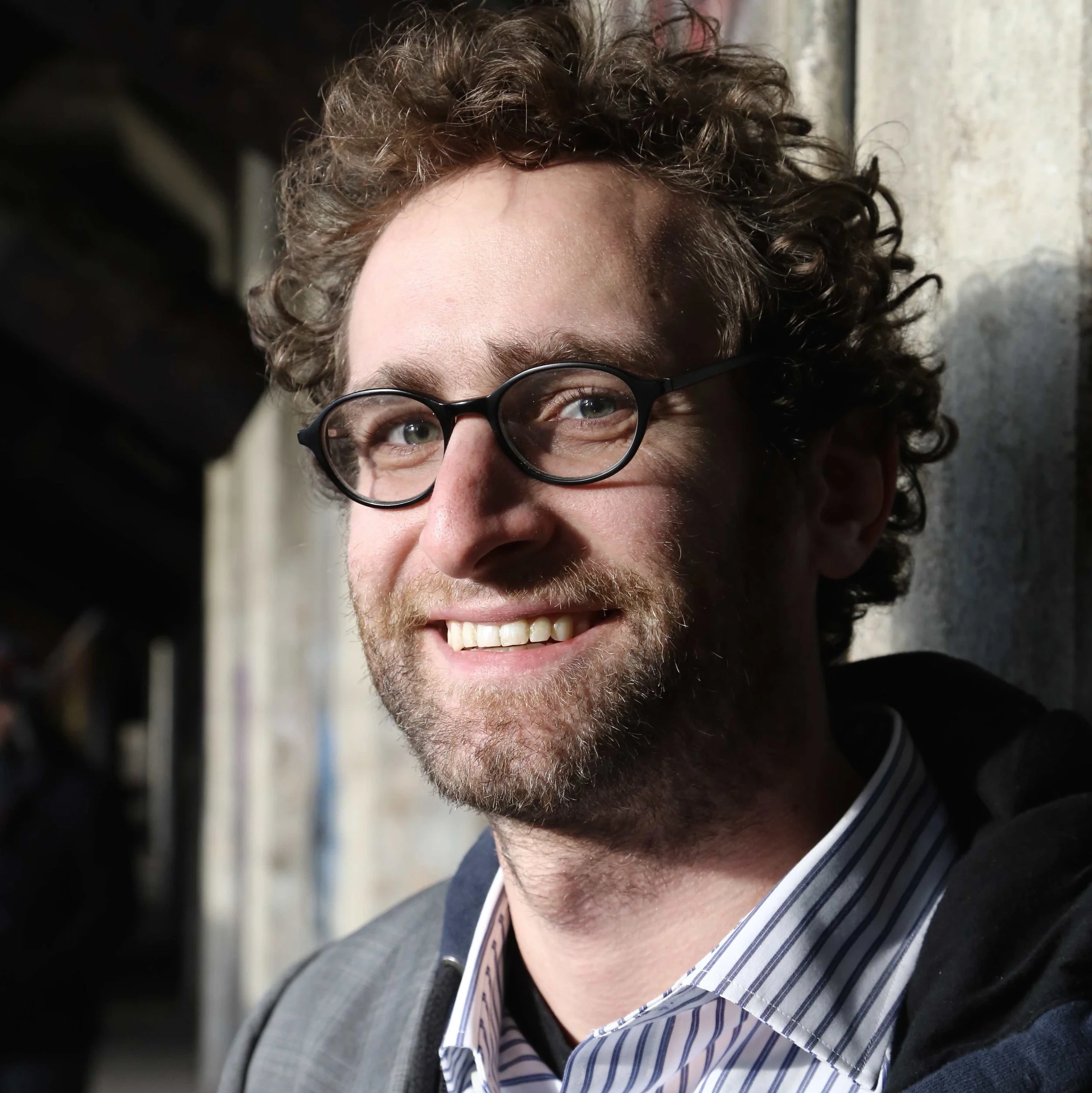 Smiling man with curly hair and glasses in a natural setting.