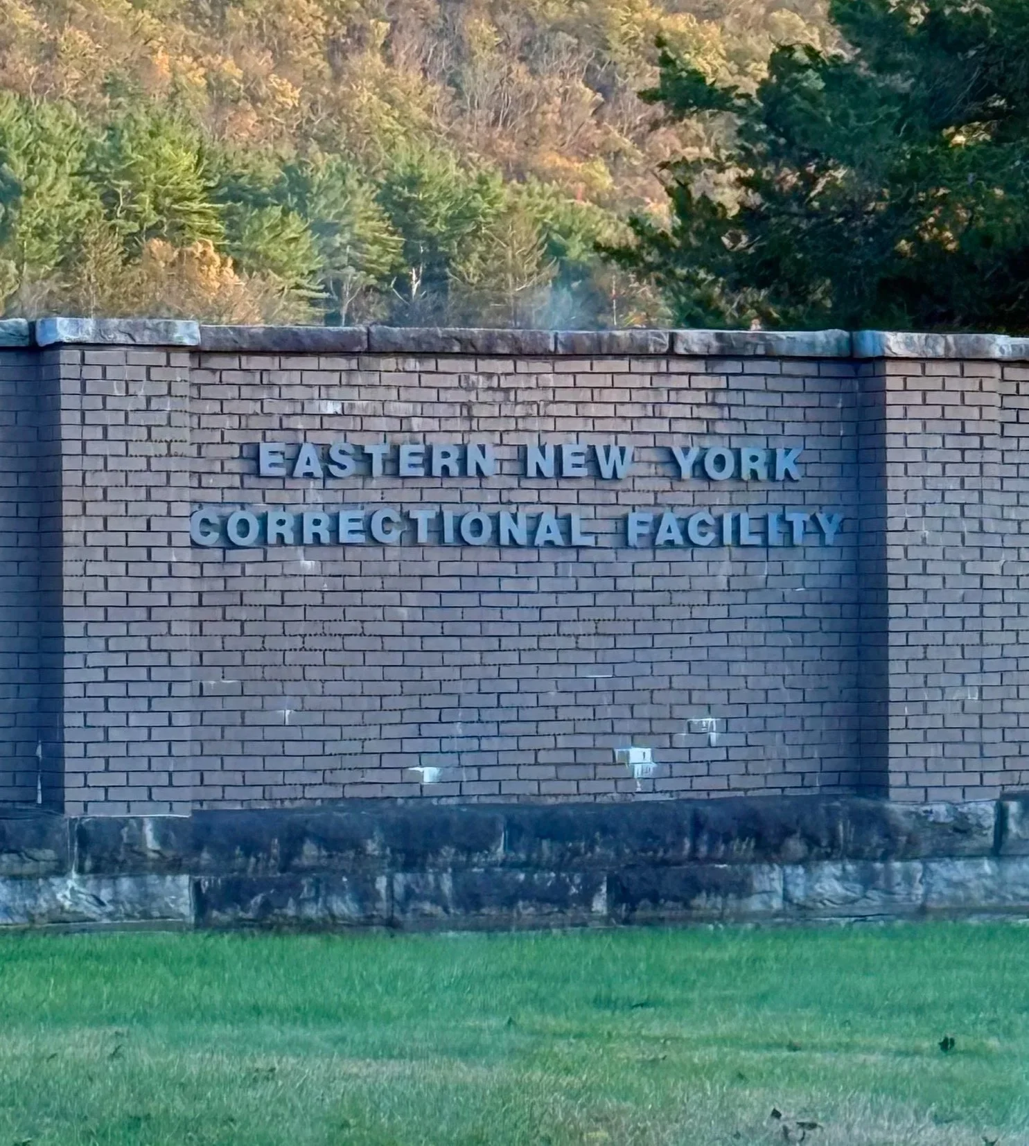 A brick wall with stone base on a grassy field with the words "Eastern New York Correctional Facility" in grey letters, and a wooded hillside behind.