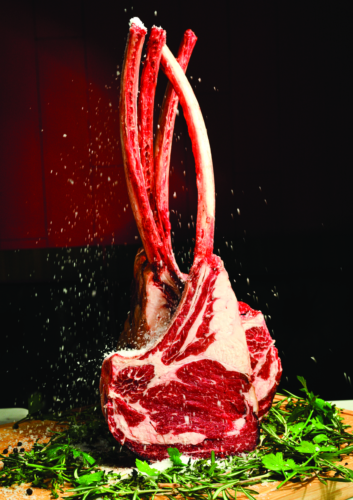 A large cut of raw beef with bones standing upright on a wooden board, with herbs and spices scattered around, against a dark background.