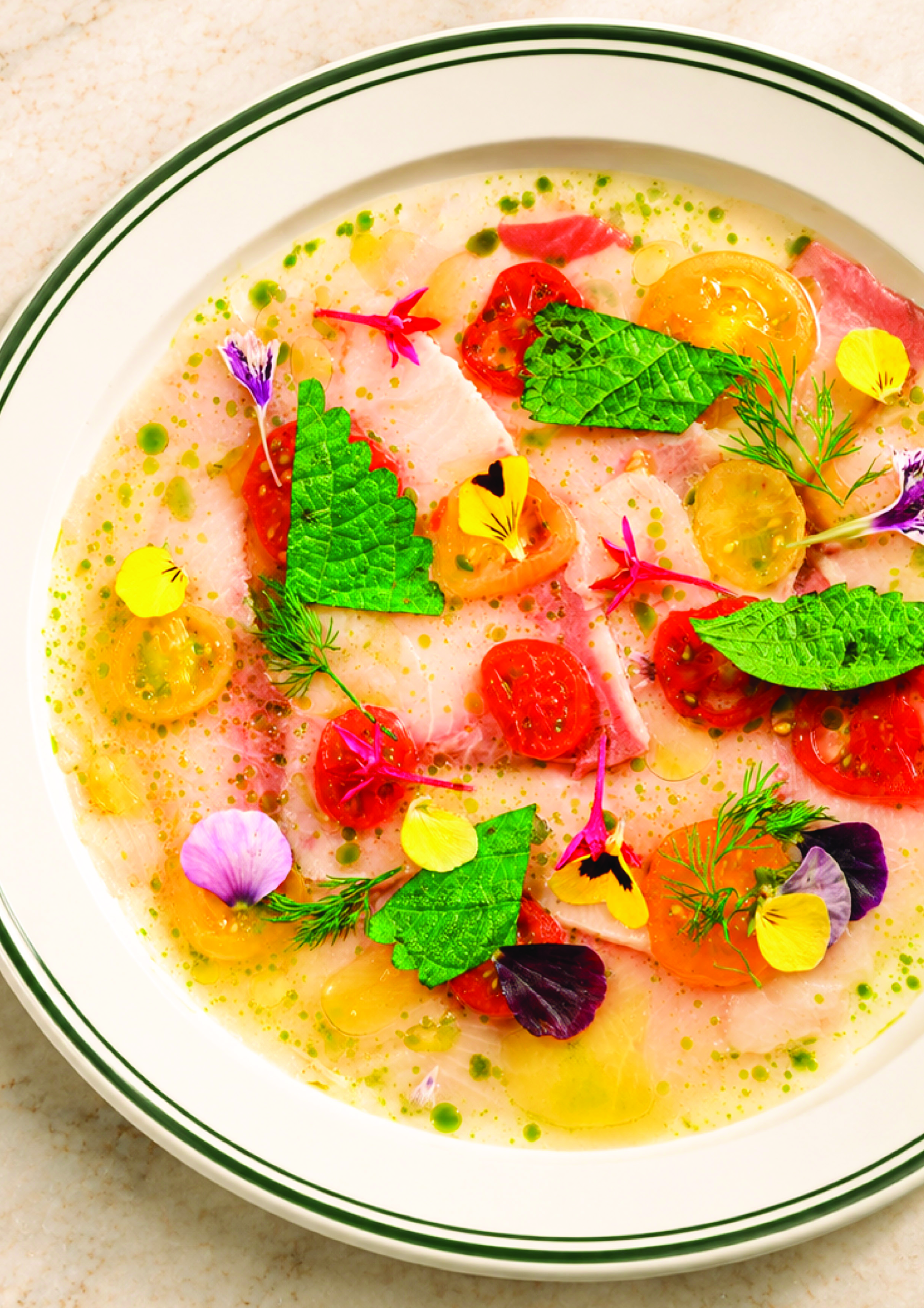 A bowl of seafood ceviche garnished with colorful edible flowers, fresh herbs, cherry tomatoes, and a drizzle of green sauce.