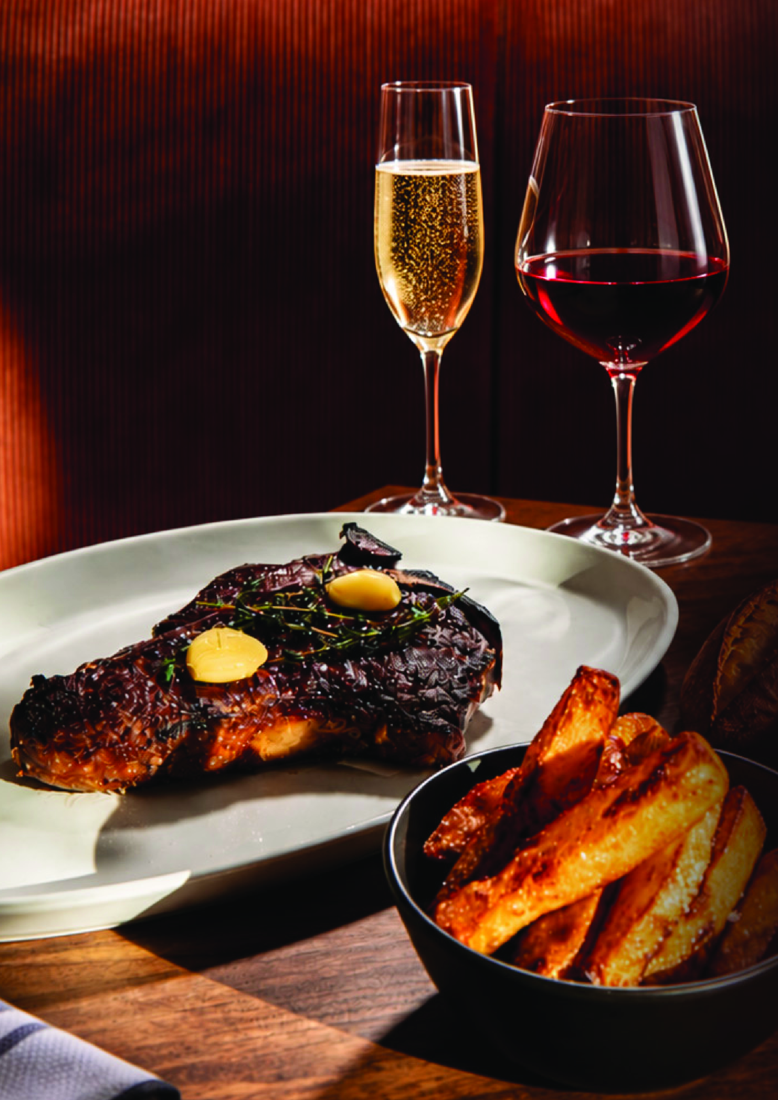 A plate with cooked meat topped with herbs, a bowl of potato wedges, a glass of champagne, and a glass of red wine on a wooden table with a dark background.