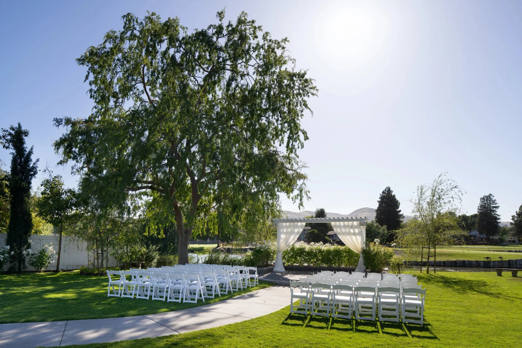 Garden wedding ceremony setting beside the pond at San Ramon Waters, with white chairs and a draped arbor