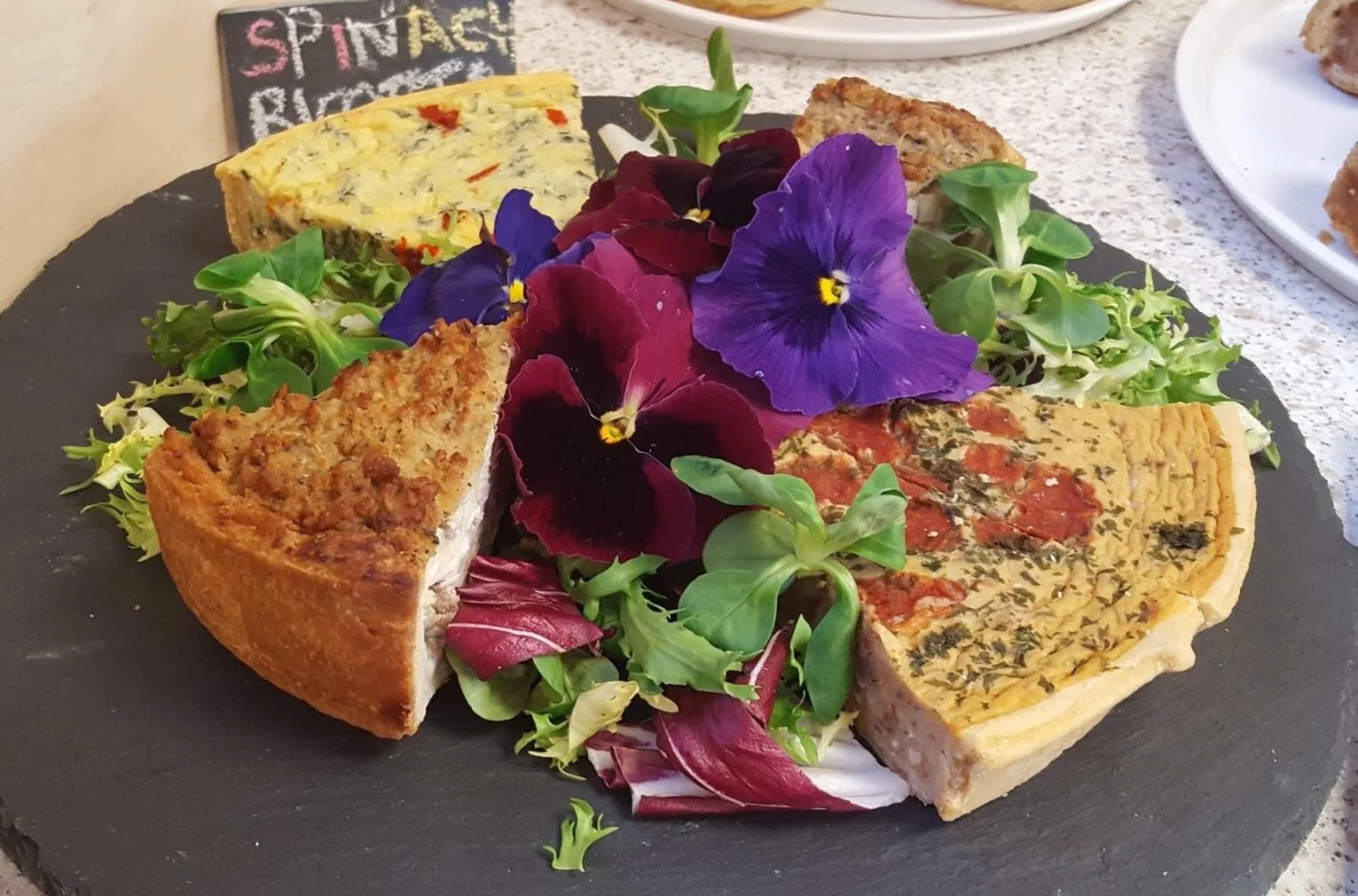 A slate with slices of quiche and tarts, garnished with salad and flowers.