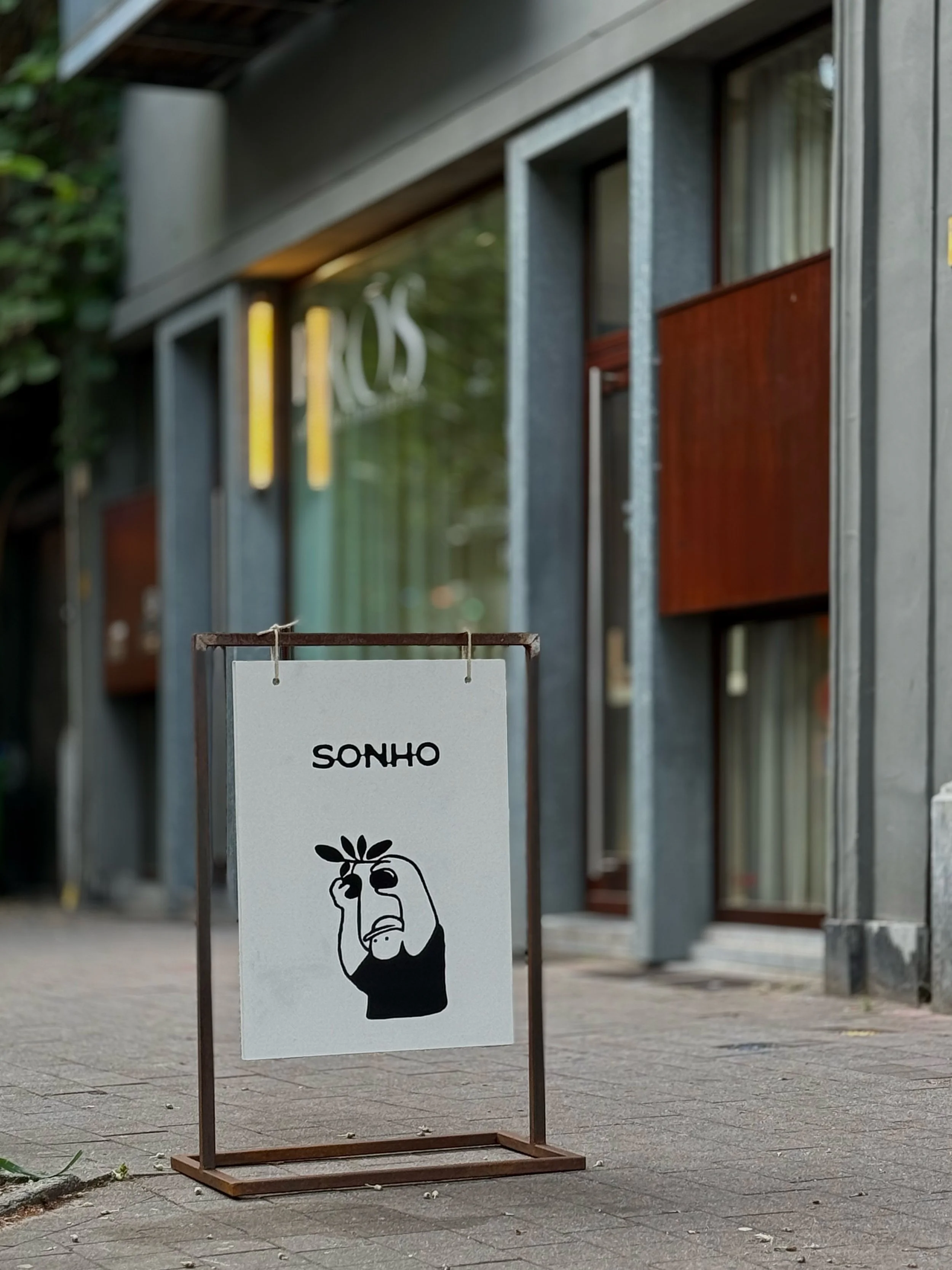 A sidewalk sign with the word 'SONHO' and a minimalist illustration of a person holding a plant over their eye, outside a modern building with large windows and metal framing.