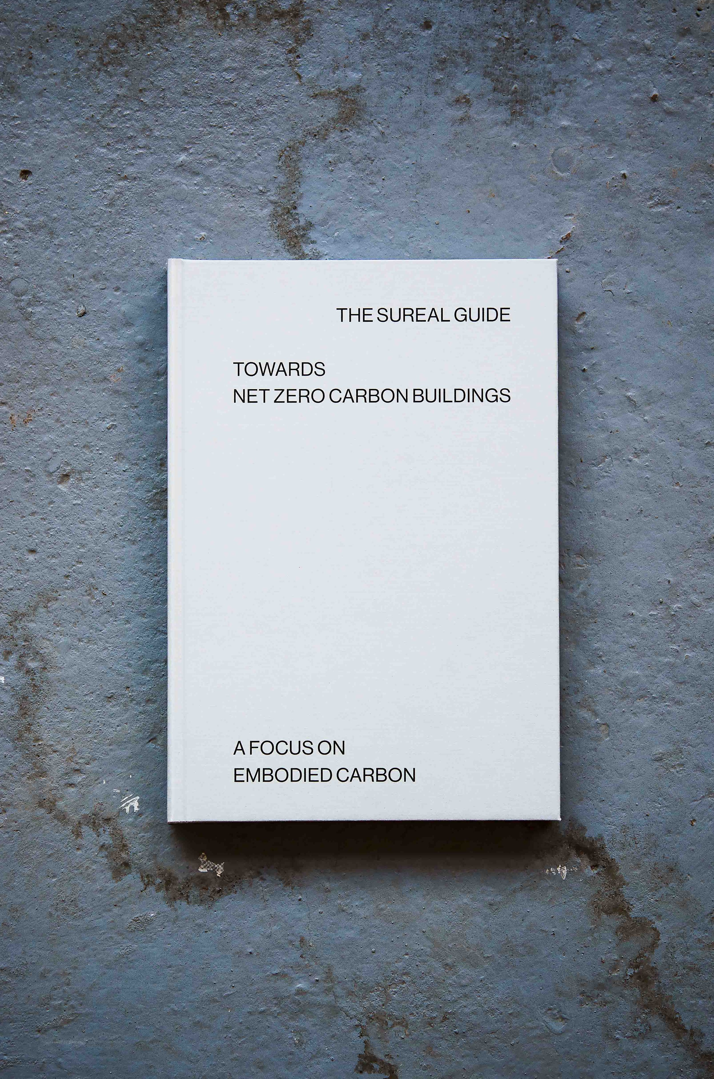 White book titled 'The Surreal Guide Towards Net Zero Carbon Buildings' on a gray concrete surface.