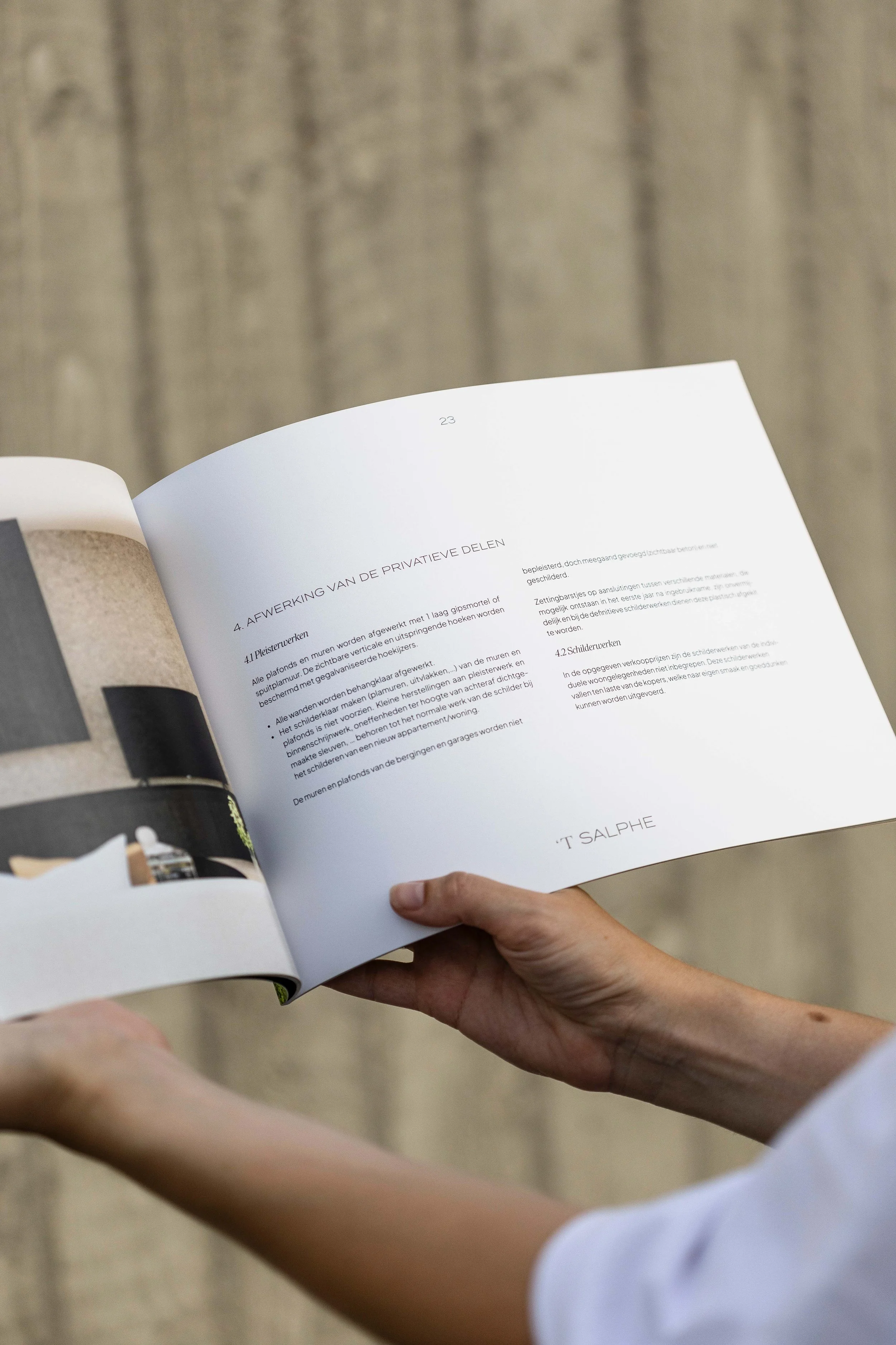 Person holding open magazine or brochure displaying pages with text and a small image, with a wooden wall background.