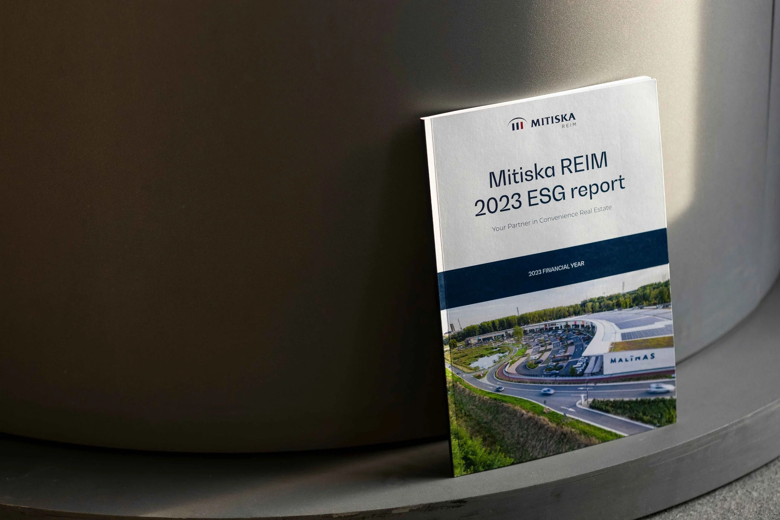 A brochure titled 'Mitiska REIM 2023 ESG report' leaning against a metallic surface, featuring a photo of a modern building with a parking lot and greenery.