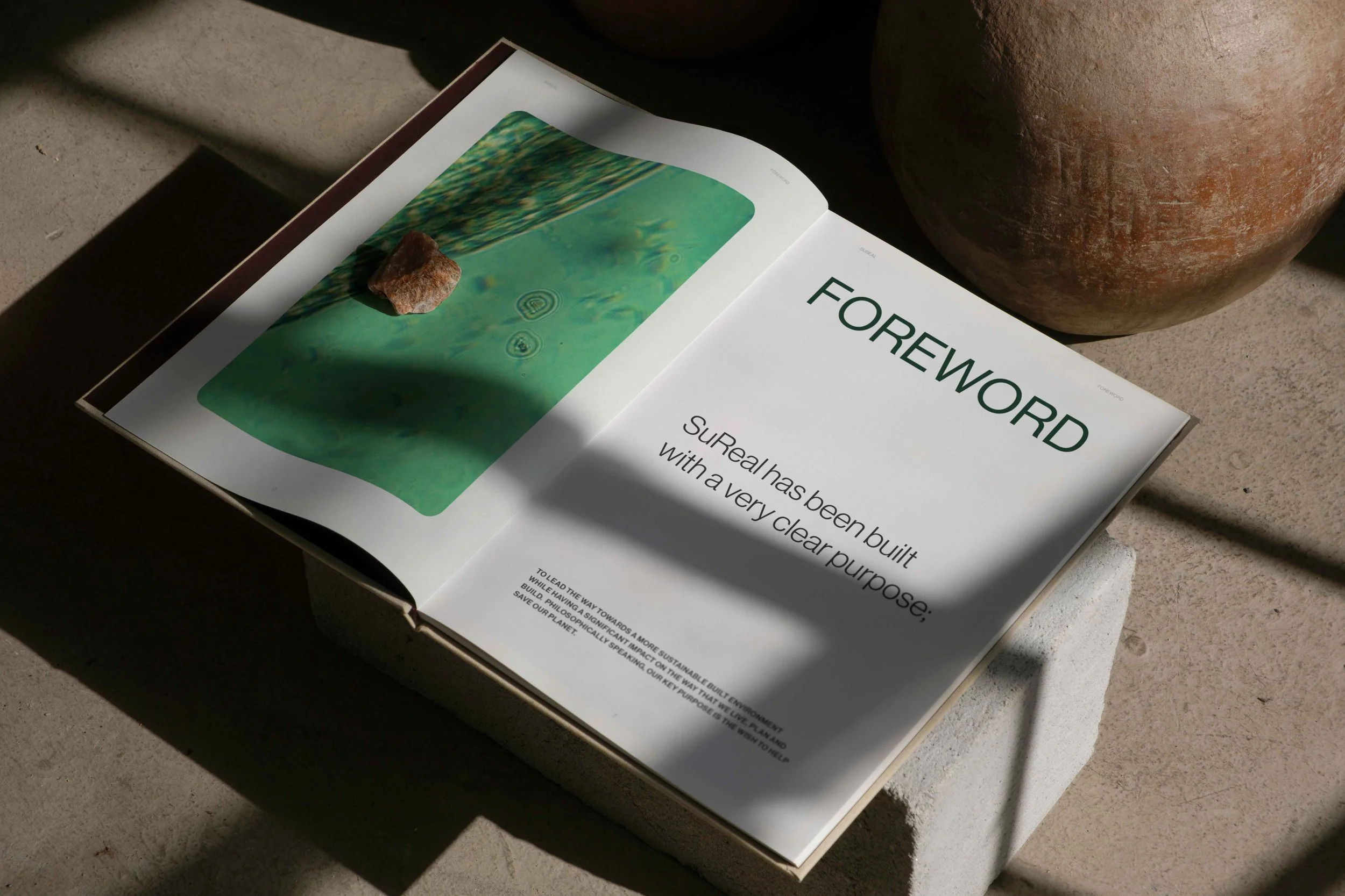 Open magazine with a white page, featuring the word "FOREWORD" in large green letters, and a paragraph of text beneath it. The magazine is placed on a concrete surface, with shadow cast across the pages, next to a large terracotta pot.
