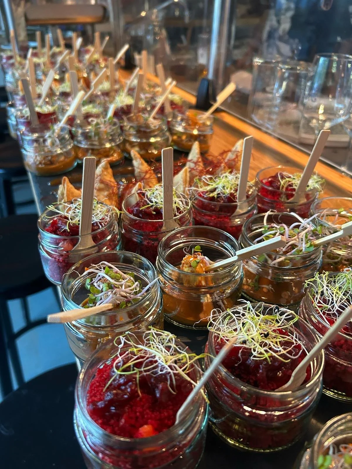 Verschiedene kleine Gläser mit bunten Vorspeisen, garniert mit Sprossen, mit kleinen Holzstäbchen als Besteck. Auf einem dunklen Tisch, im Hintergrund ein Barbereich. Event Catering Wiesbaden