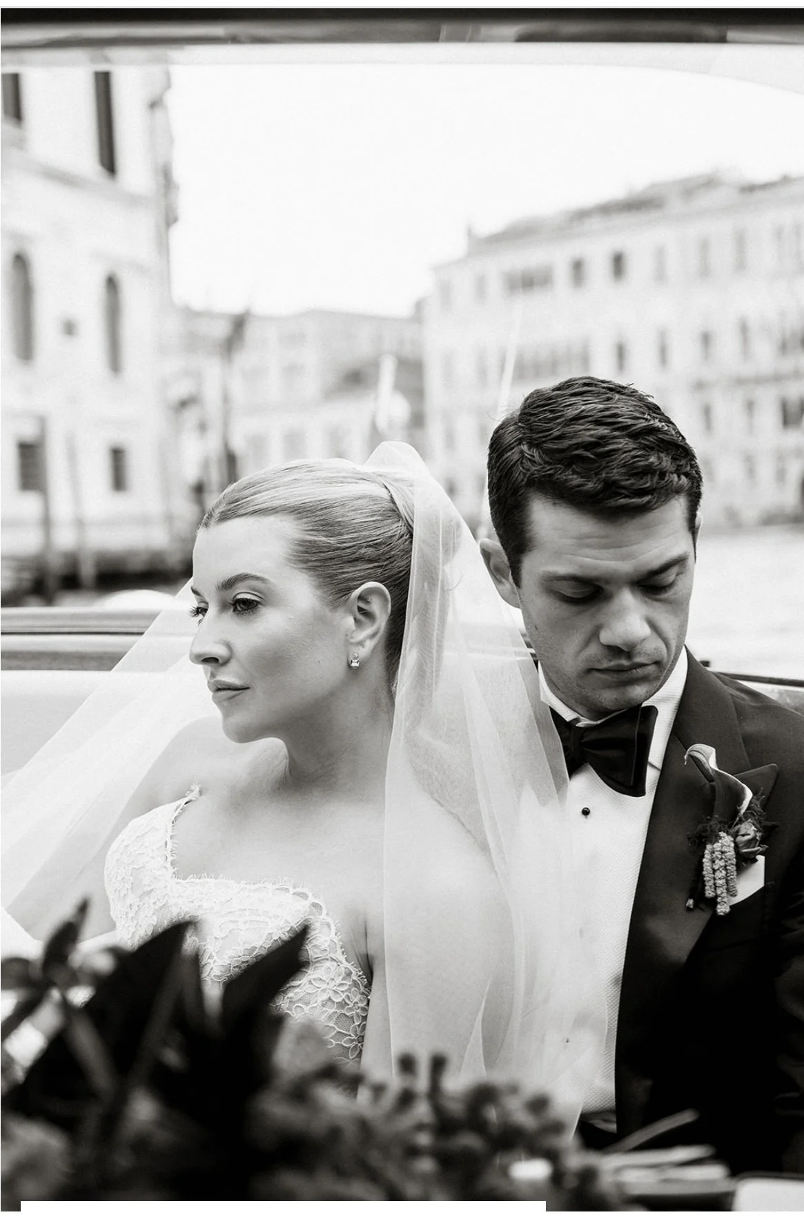 Black and white photo of a bride and groom sitting in a vintage convertible car with a cityscape background. The bride has her hair up with a veil, and the groom is in a tuxedo with a boutonniere.