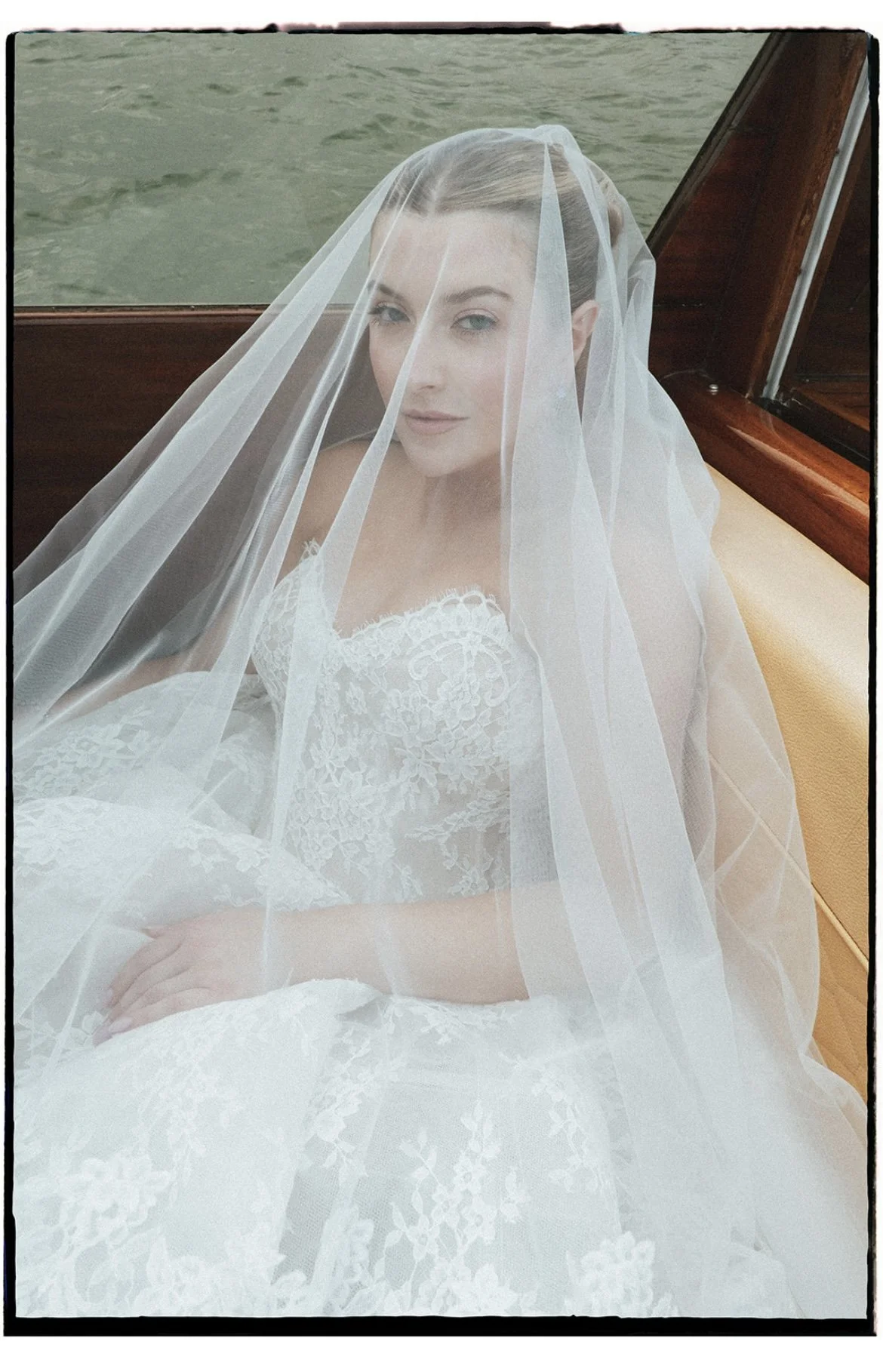 A bride sitting on a boat with a veil over her face, wearing a white lace wedding dress, with water in the background.