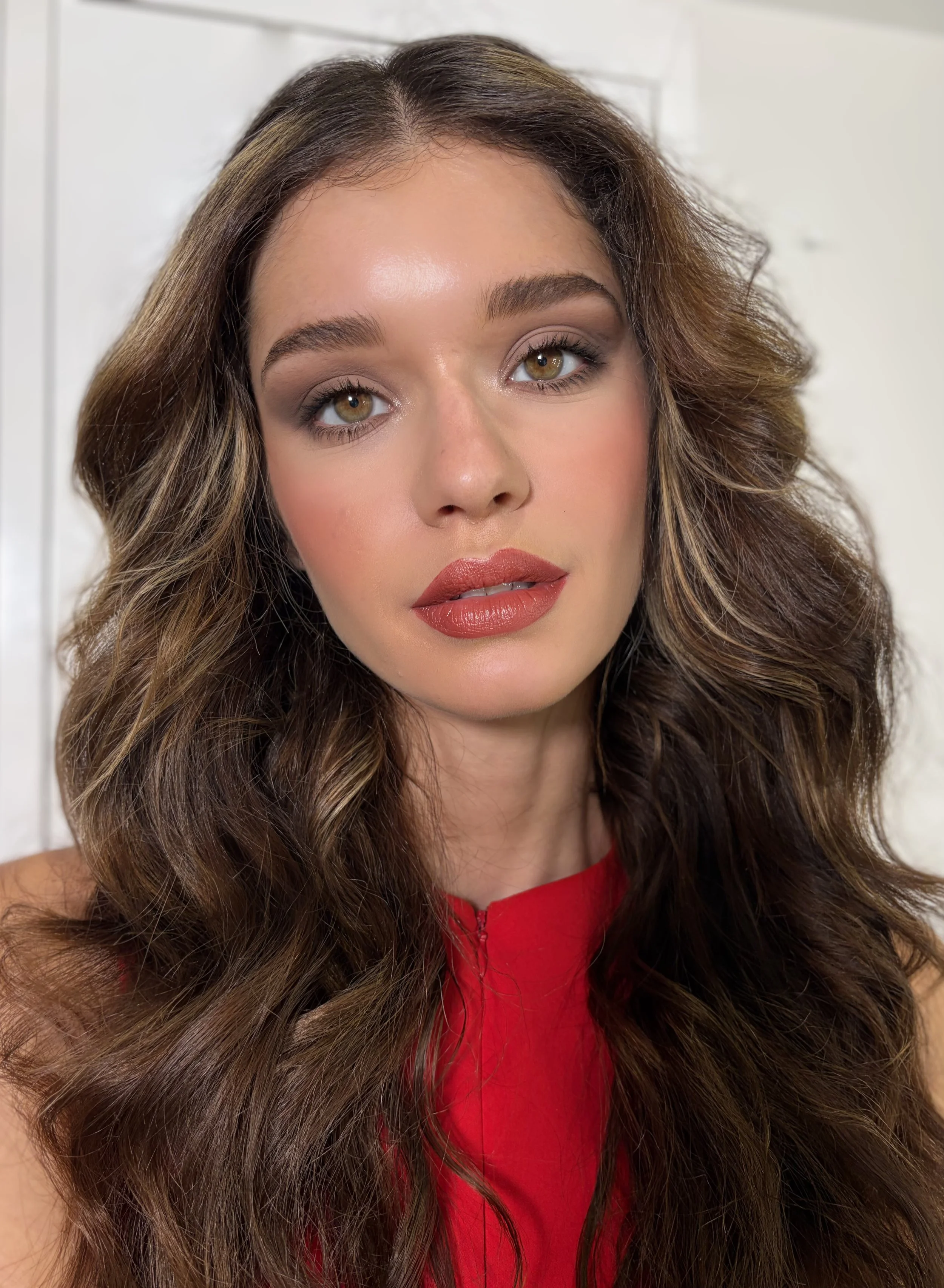 Close-up of a woman with long, wavy brown hair, wearing makeup, and a red top, looking directly at the camera.
