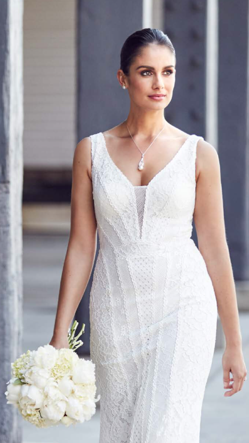 A woman in a white lace wedding dress holding a bouquet of white flowers, standing outdoors with a neutral background.