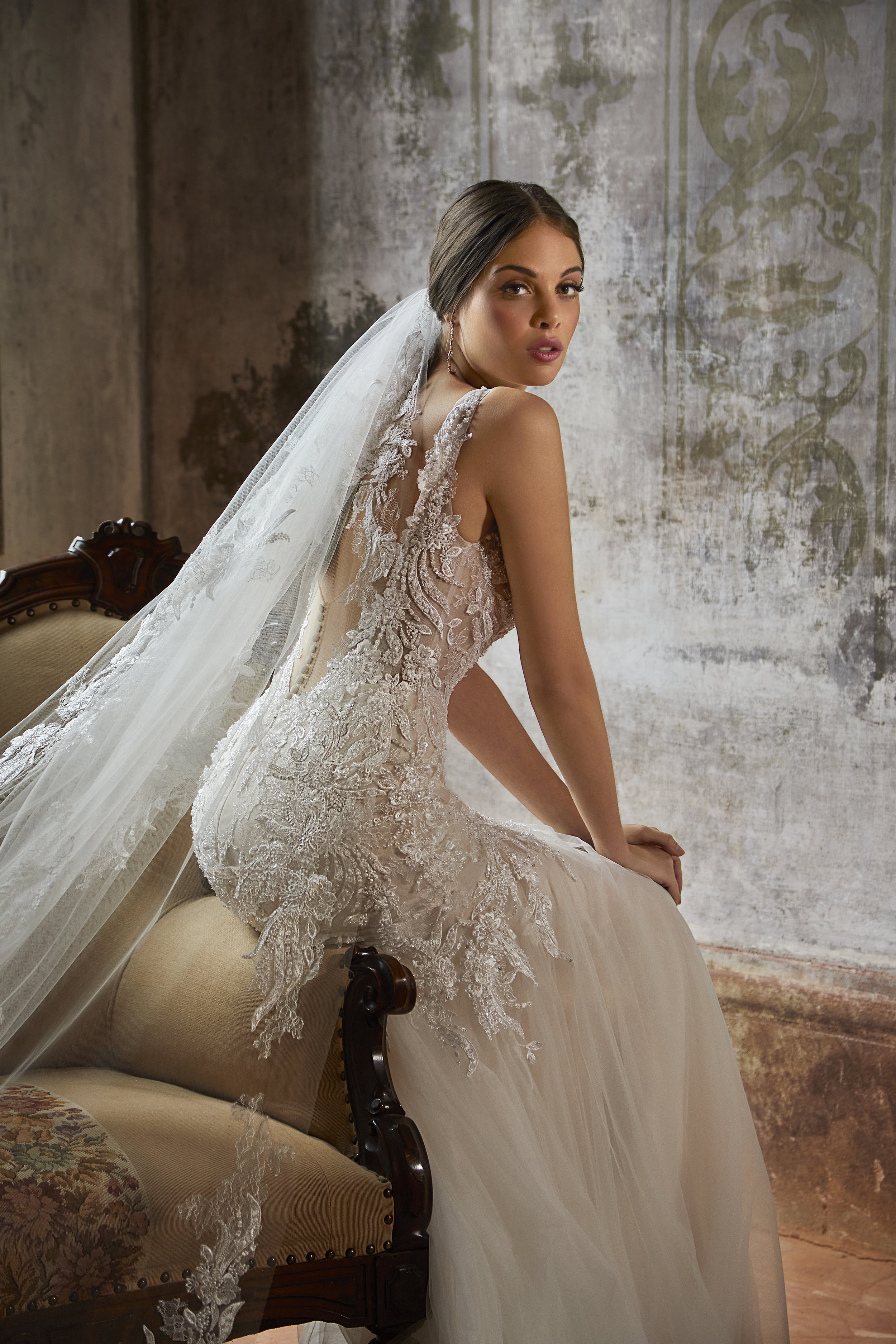 A woman dressed in a wedding gown with lace details and a veil, sitting on a vintage-style sofa in an old room with aged walls and decorative patterns.