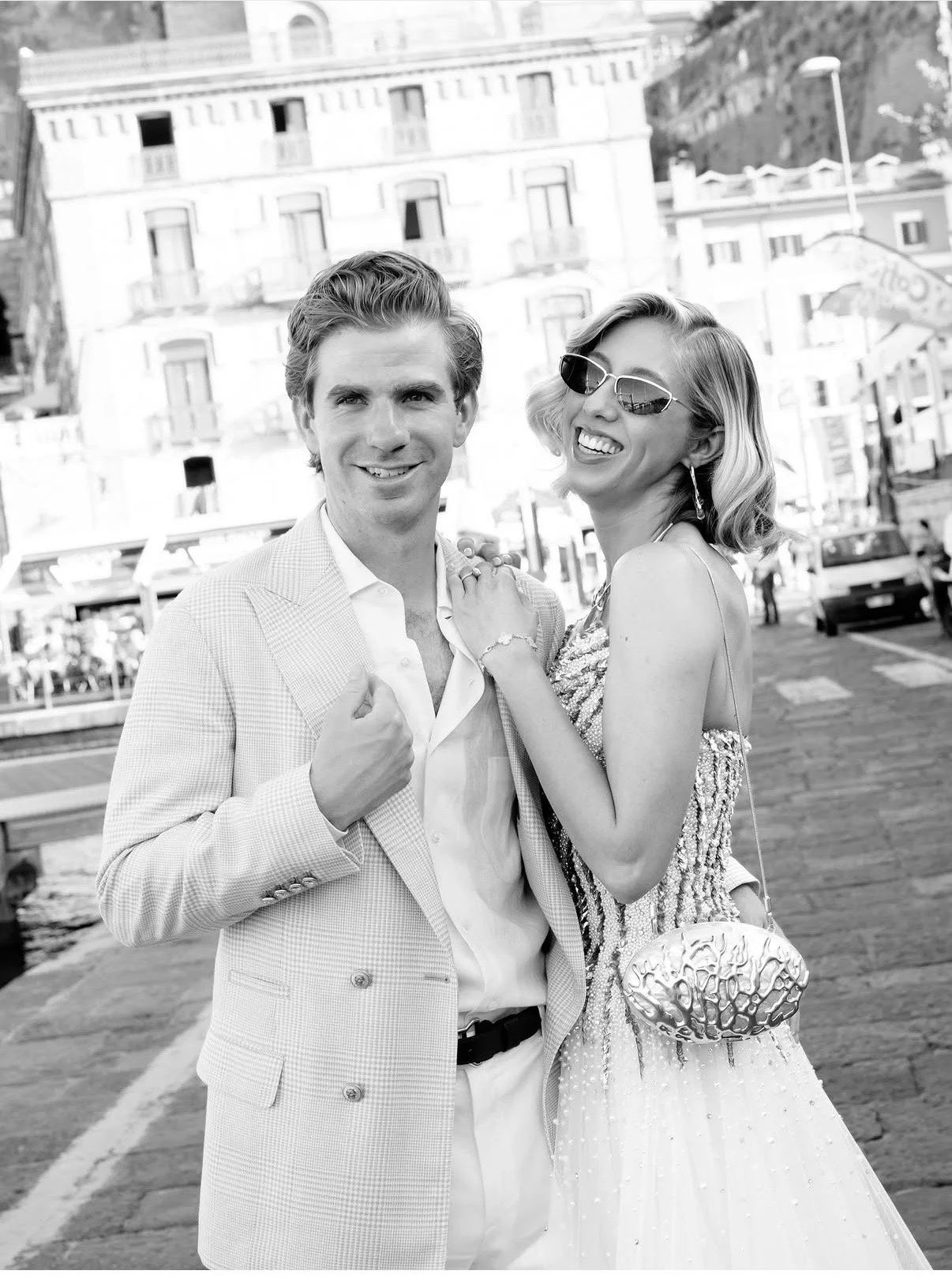 A smiling man in a light-colored suit with a checked pattern, and a woman wearing sunglasses, a sparkling dress, and jewelry, standing outdoors near a waterfront with buildings and cars in the background.