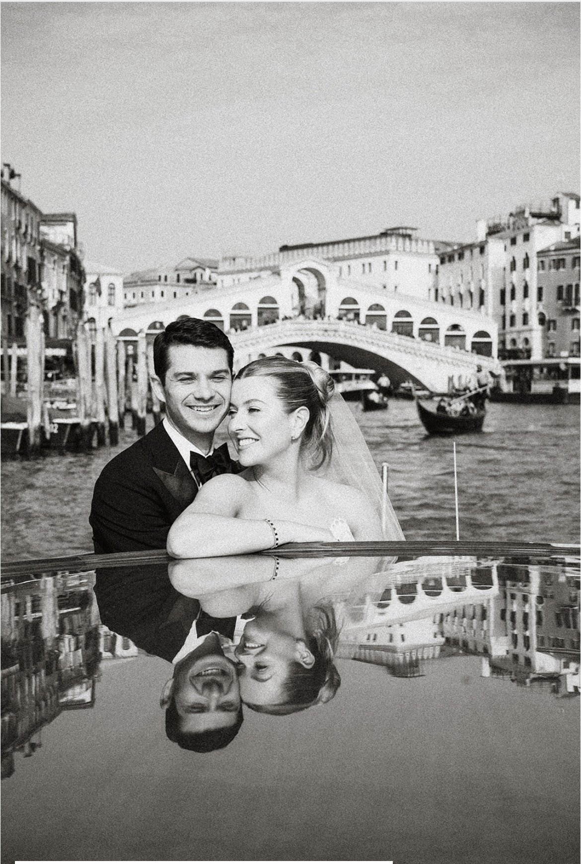 Black and white photo of a newlywed couple smiling and posing with Venice's Grand Canal and Rialto Bridge in the background.