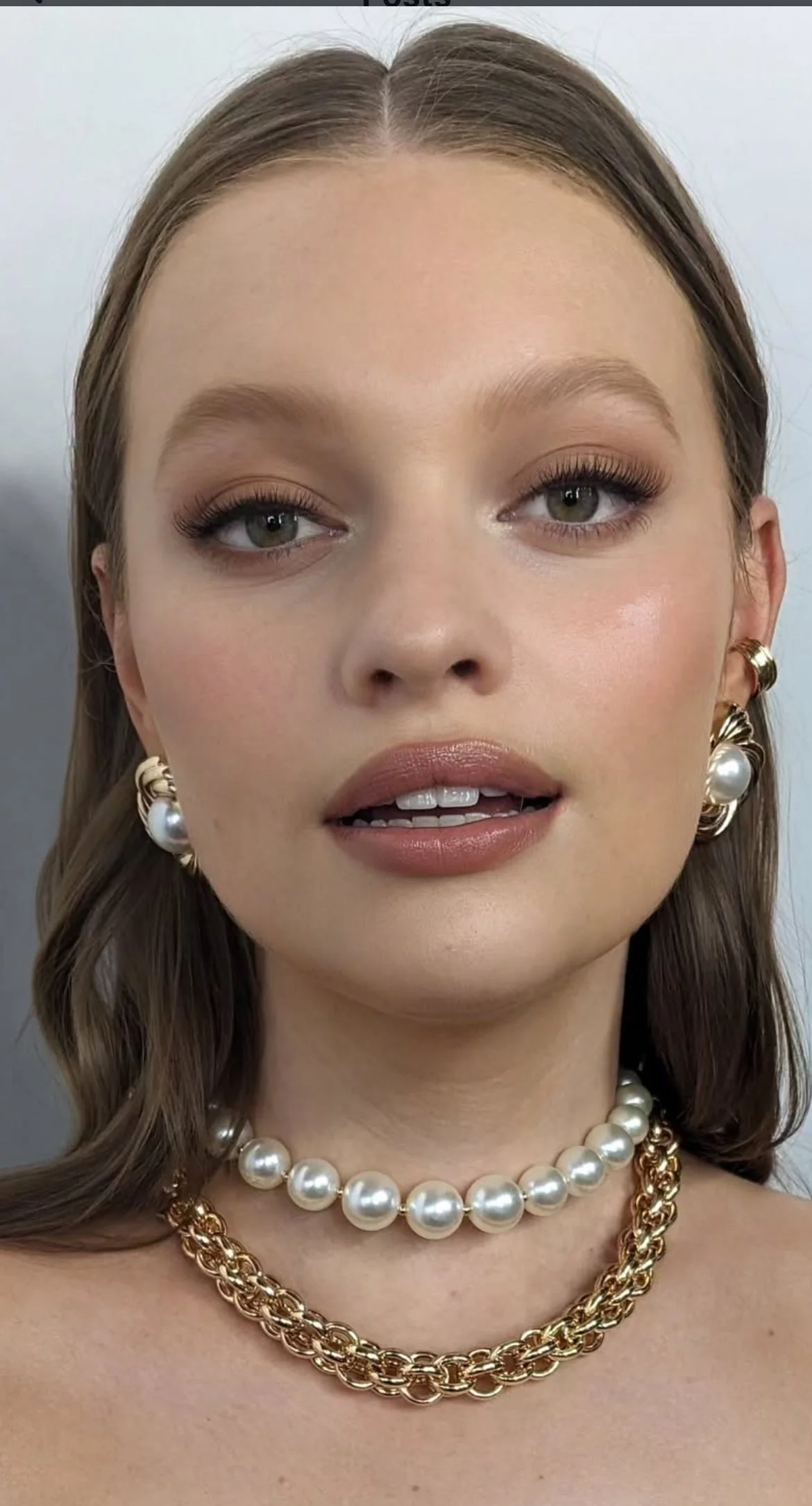 A woman with light skin and gray eyes wearing pearl earrings, a pearl necklace, and a gold chain necklace, with makeup and styled hair.