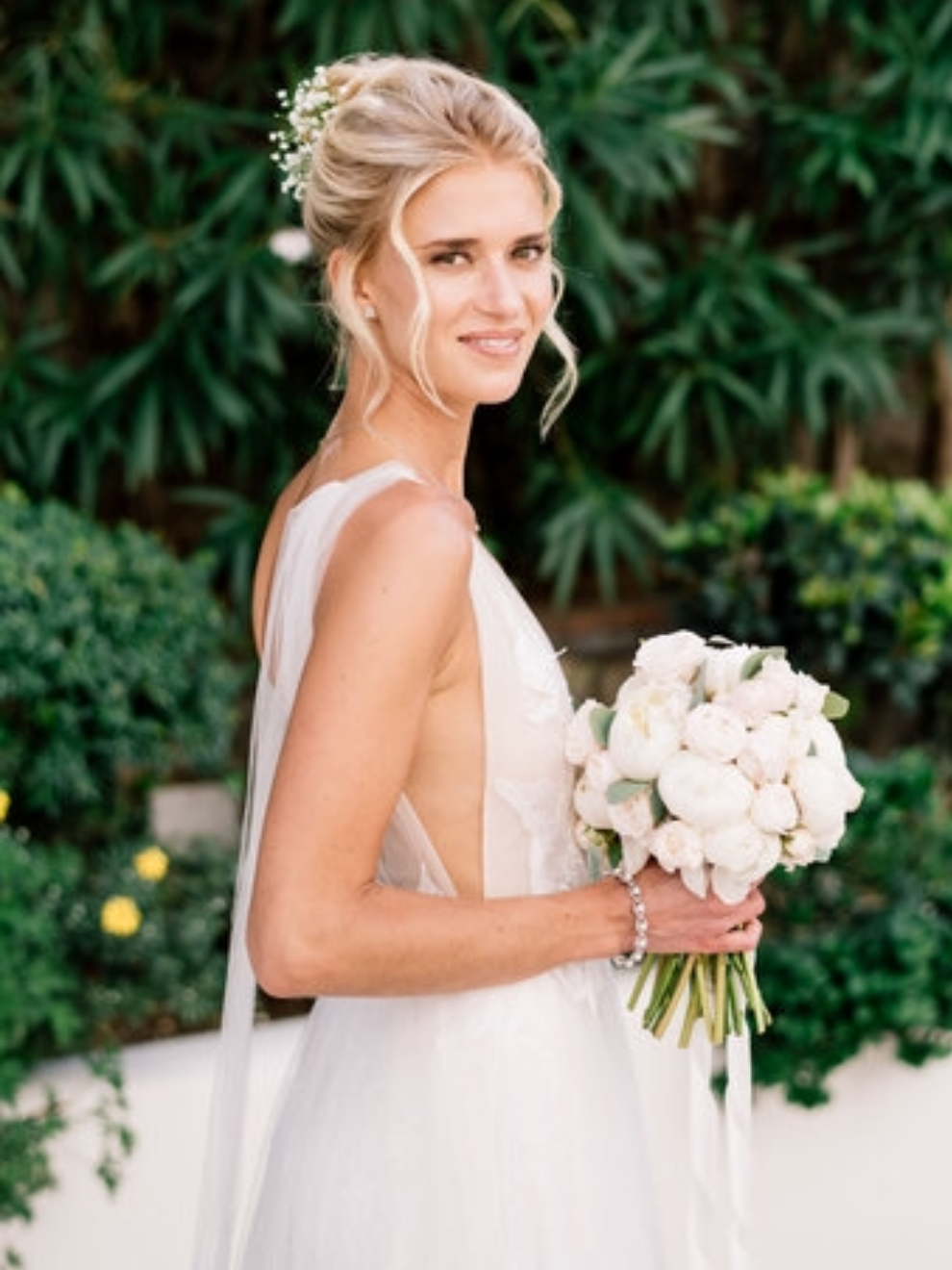 A smiling bride with blonde hair in an elegant updo, holding a bouquet of white roses, wearing a sleeveless white wedding dress, standing outdoors with green foliage background.