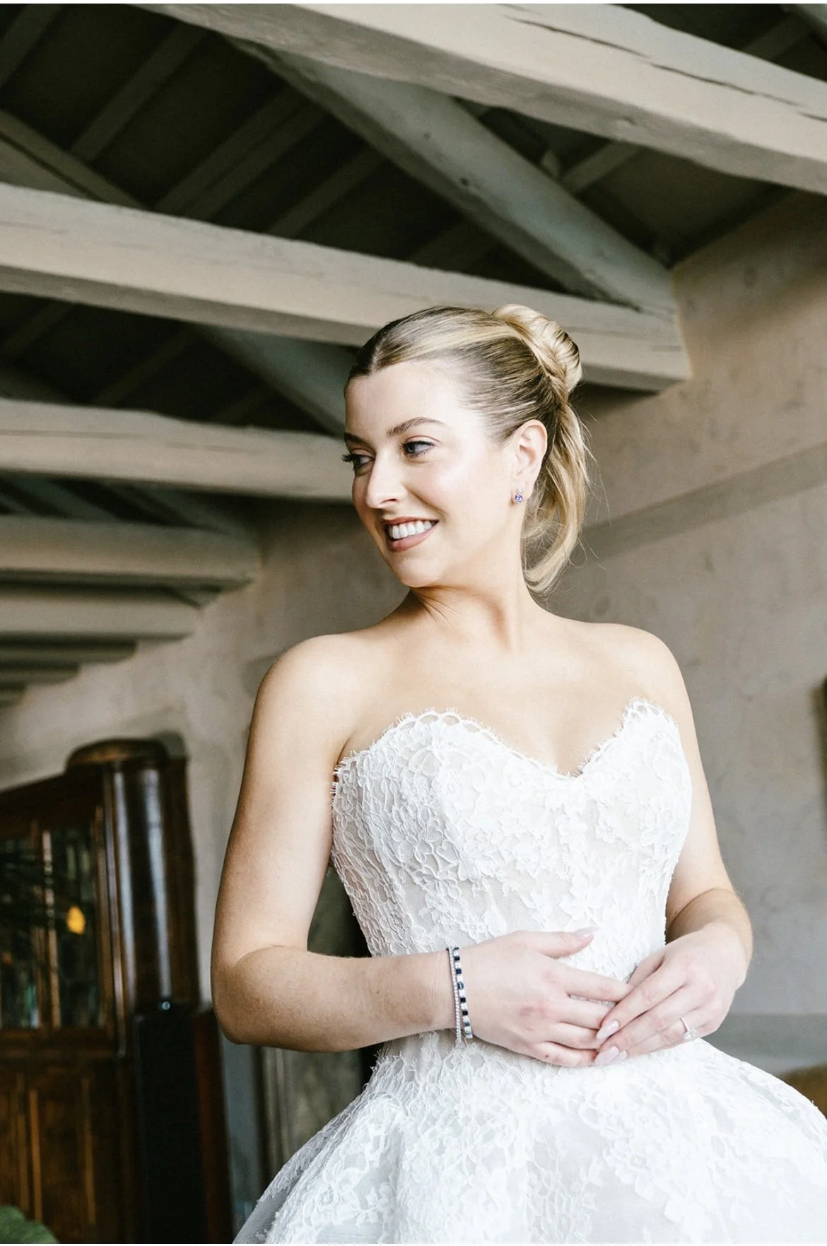 A woman in a white lace wedding dress, smiling, with her hair styled in an updo, standing in a room with wooden beams overhead and furniture in the background.