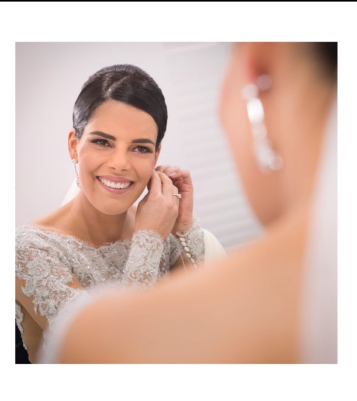 A bride with short dark hair in a lace wedding dress is smiling while adjusting her earring, looking at herself in a mirror.