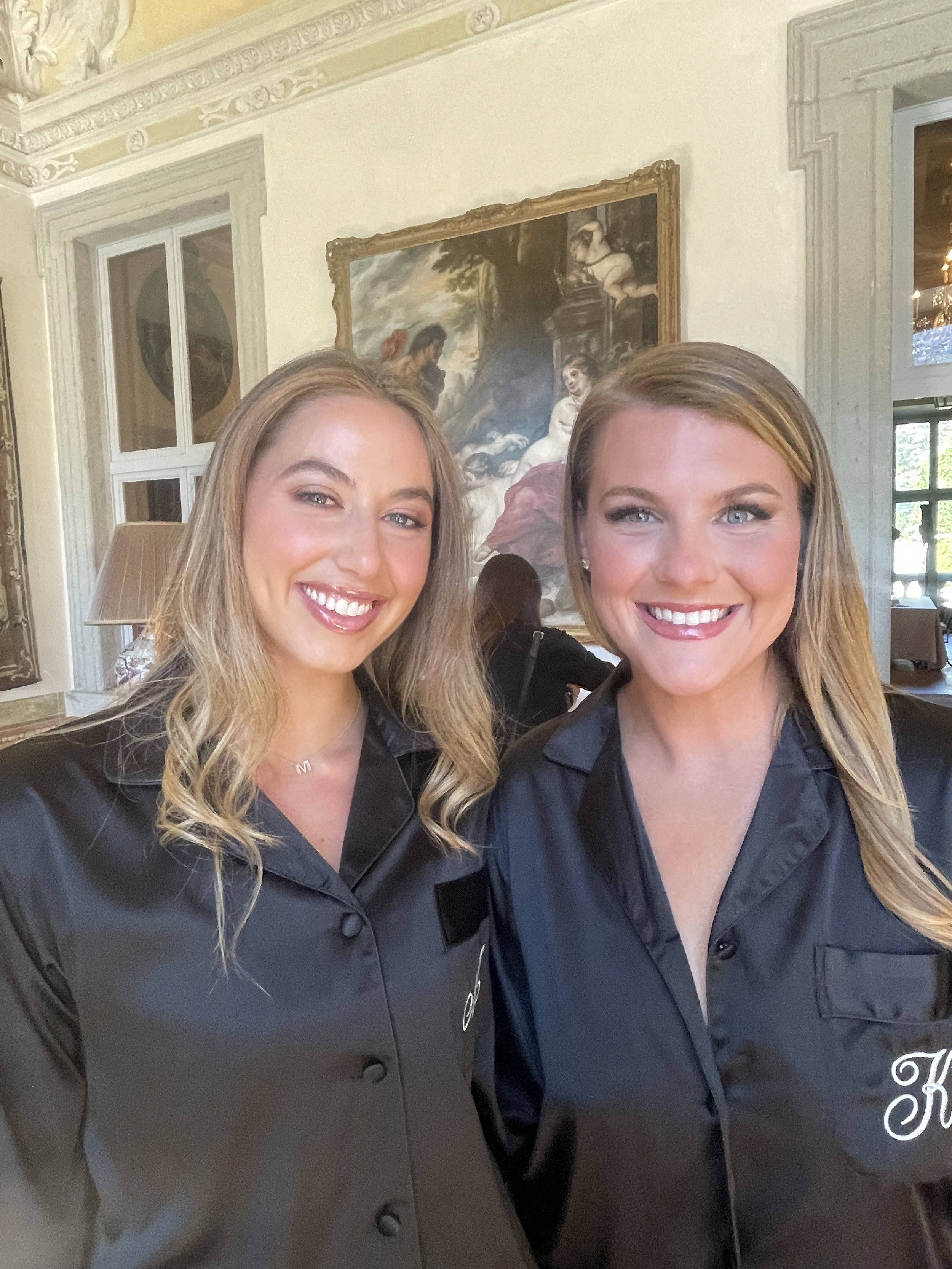 Two women smiling and taking a selfie together in a room with ornate decoration, a large painting, and a window. Both women are wearing dark shirts.