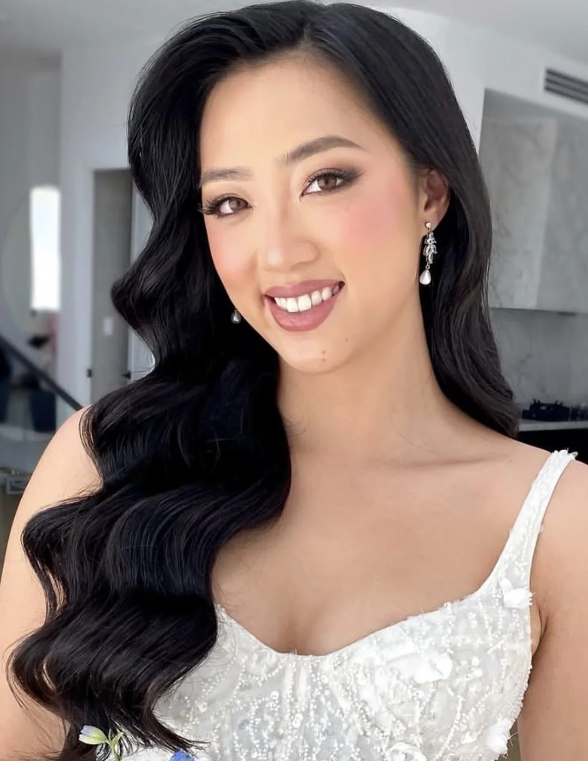 A young woman with long, wavy black hair smiling, wearing earrings and a white dress with floral details, indoors with a modern background.