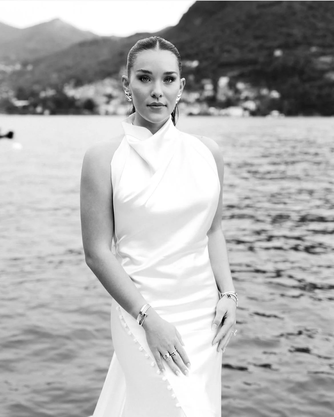 A woman in a satin dress standing by a body of water with mountains and houses in the background.