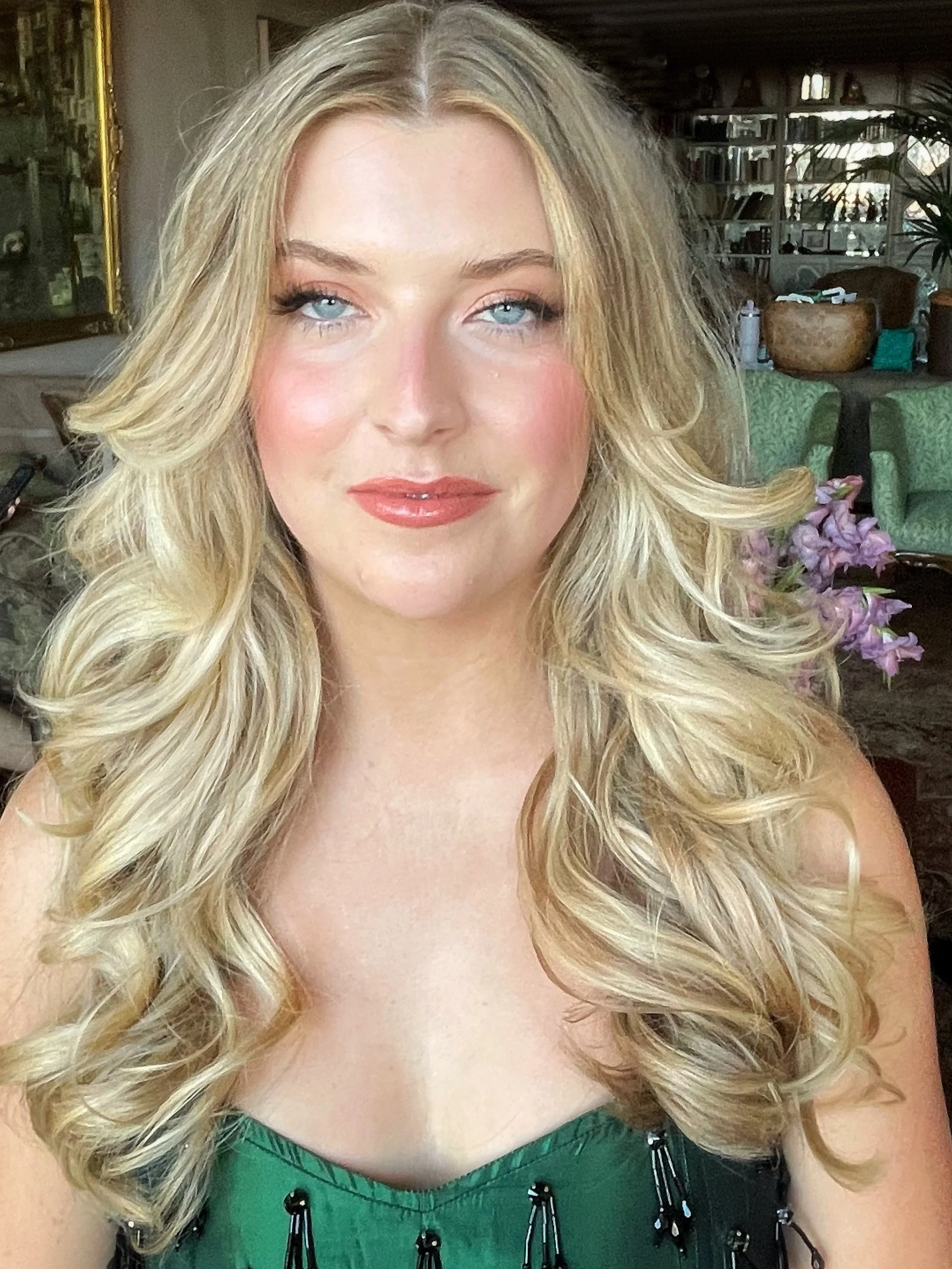 A woman with long, curly blonde hair, blue eyes, and light makeup wearing a green top with black detailing, posing indoors in front of a bookshelf and a floral arrangement.