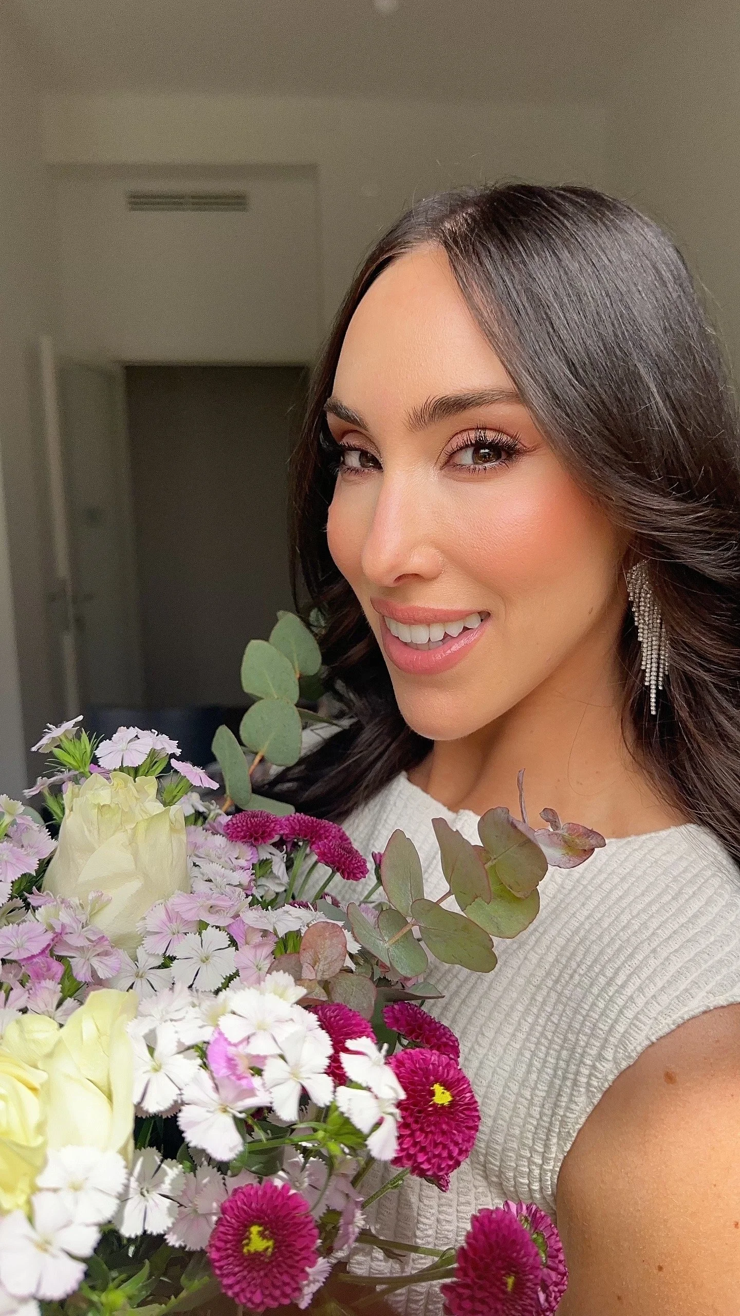 A woman smiling and holding a large bouquet of flowers, including pink, white, and purple blooms, with earrings and a white top, indoors.