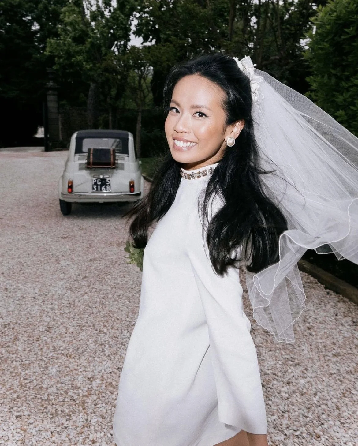 A smiling woman in a white dress with a veil indoors, with a vintage car in the background.