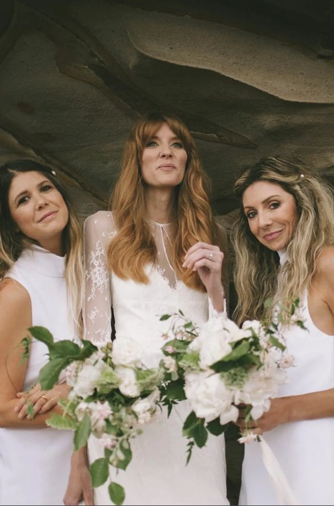 Three women dressed in white, standing under a cave-like rock formation, holding a large bouquet of white and pink flowers.