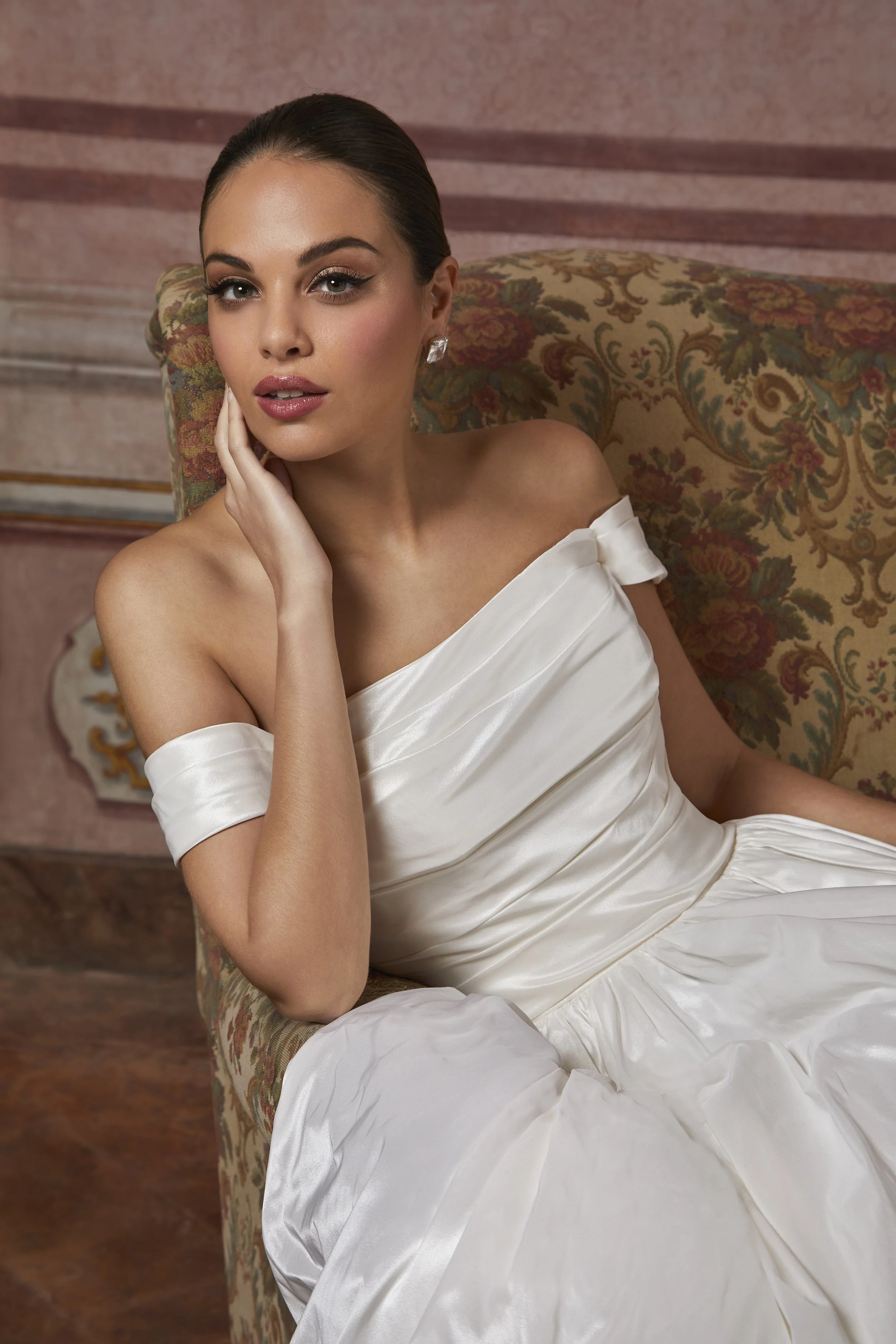 A woman in a white satin off-shoulder dress sitting on a vintage floral upholstered armchair with a pink stucco wall in the background.