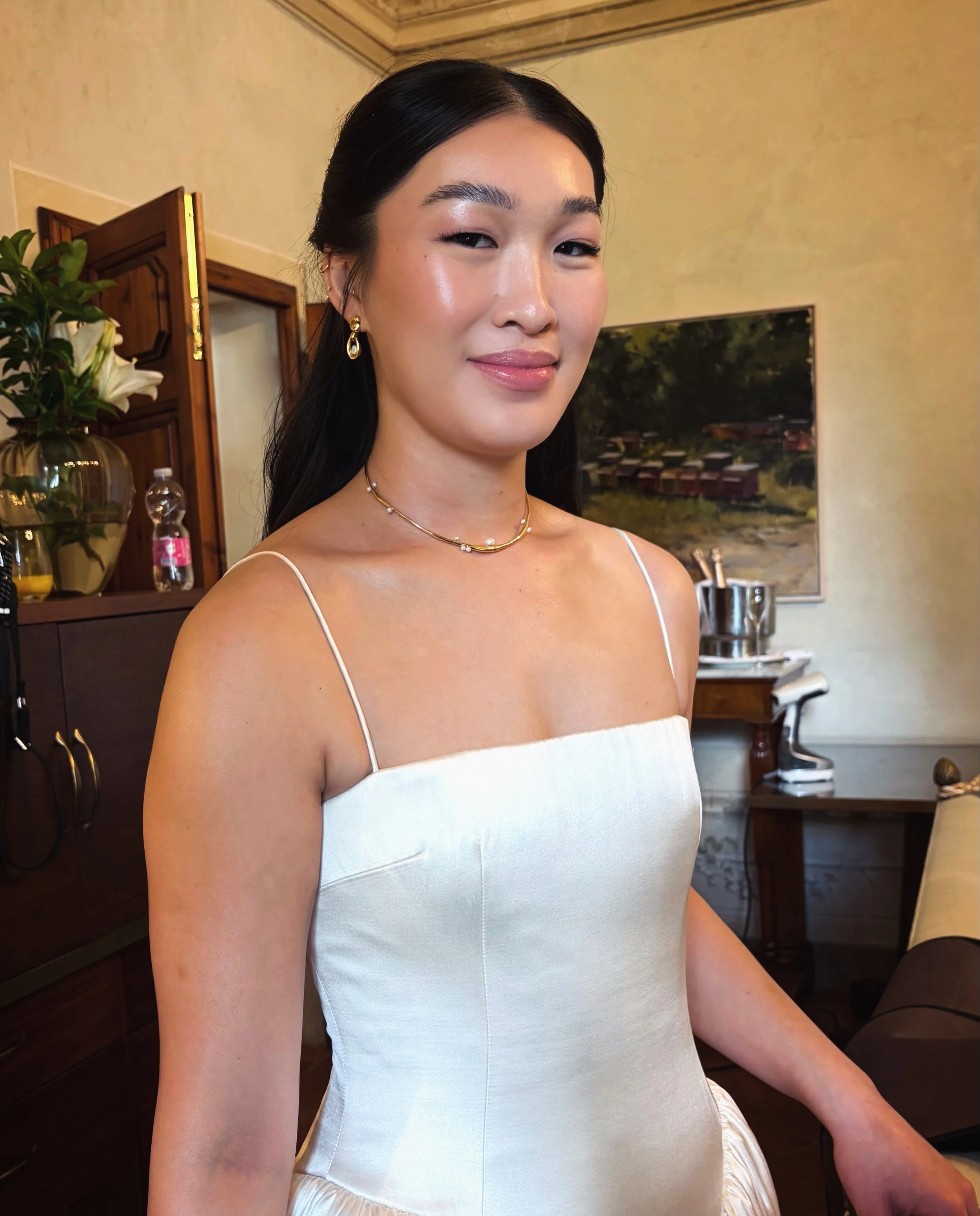 A woman dressed in a white strapless dress with jewelry, standing indoors in front of a landscape painting and other furniture.