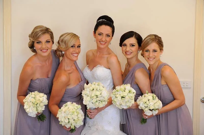 A bride in a white wedding dress standing with five bridesmaids in lavender dresses, each holding a bouquet of white flowers, inside a room with plain walls.