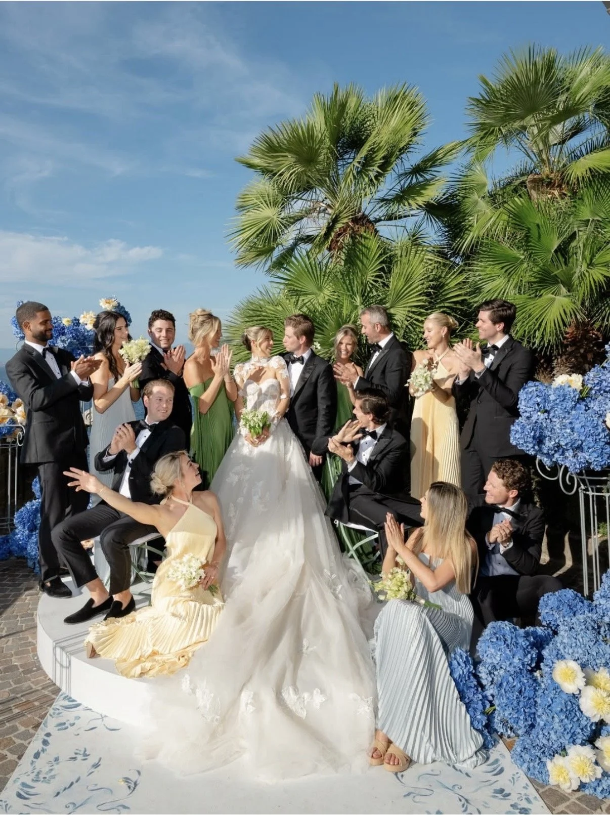 Group of people at a wedding celebration with palm trees and blue flowers in the background.