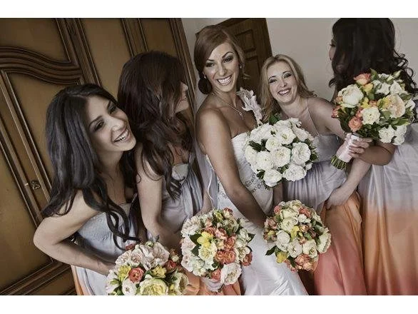 Group of five women in bridesmaid dresses holding bouquets, smiling and celebrating in an indoor setting.