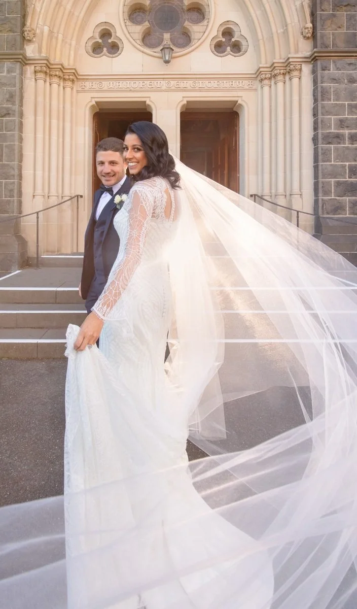 A bride and groom standing outside a church or chapel, smiling. The bride is in a white lace wedding dress with a long veil, and the groom is in a dark tuxedo with a white shirt and bow tie, in front of an ornate stone building.