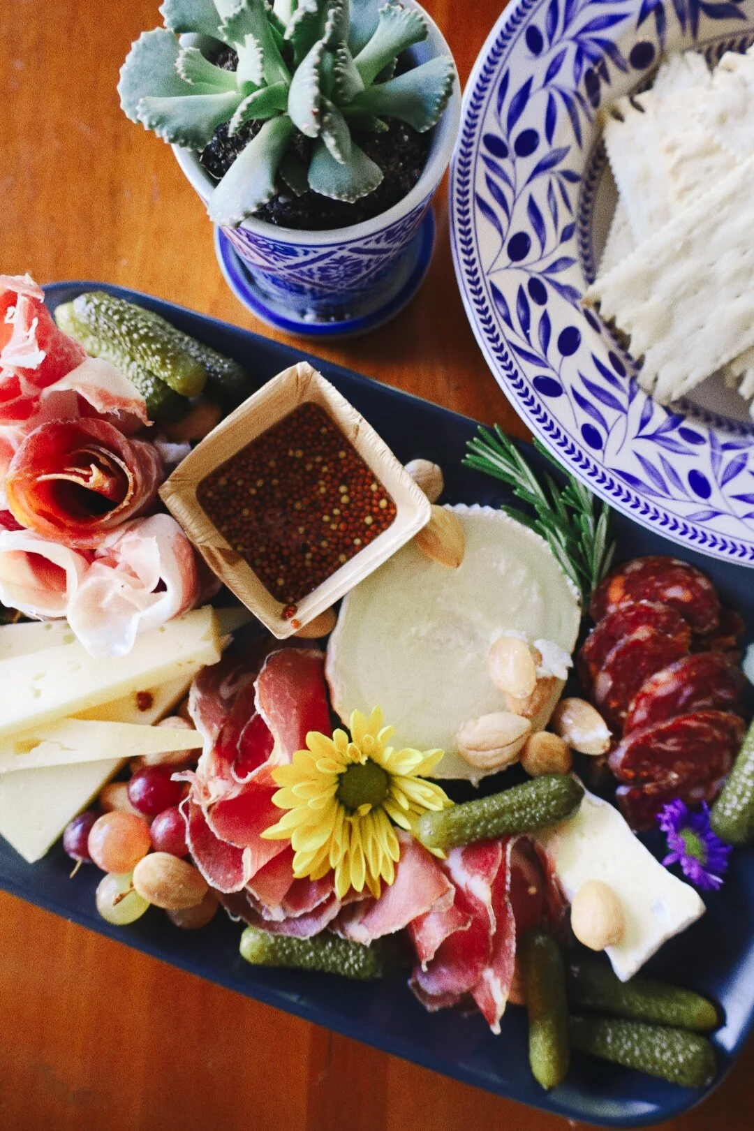 Charcuterie board with cheeses, cured meats, grapes, pickles, mustard, and garnished with flowers, next to a potted succulent and a blue patterned plate of crackers.