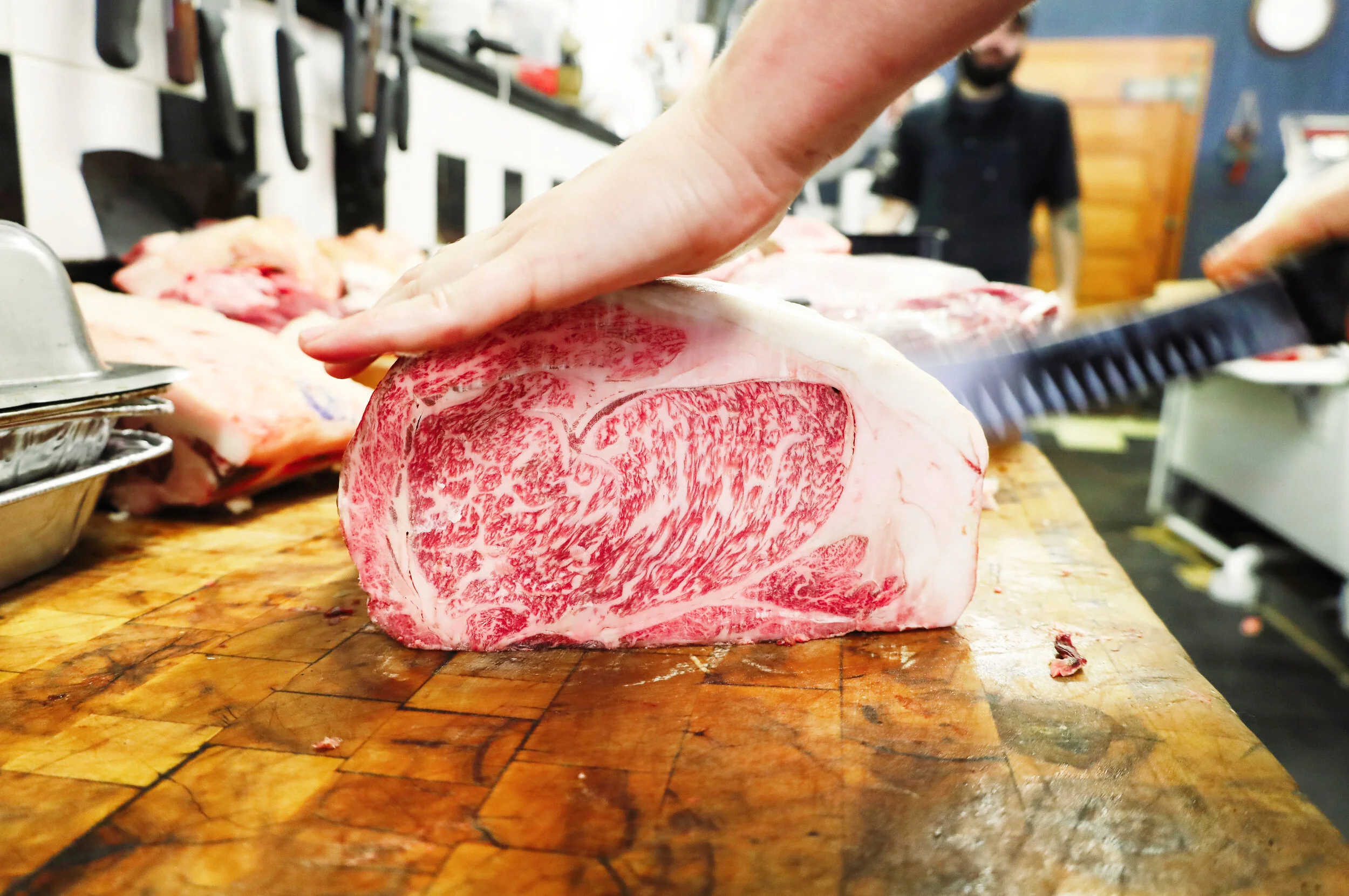 Close-up of a hand slicing a large piece of wagyu beef on a butcher's block, with a person in black in the background.