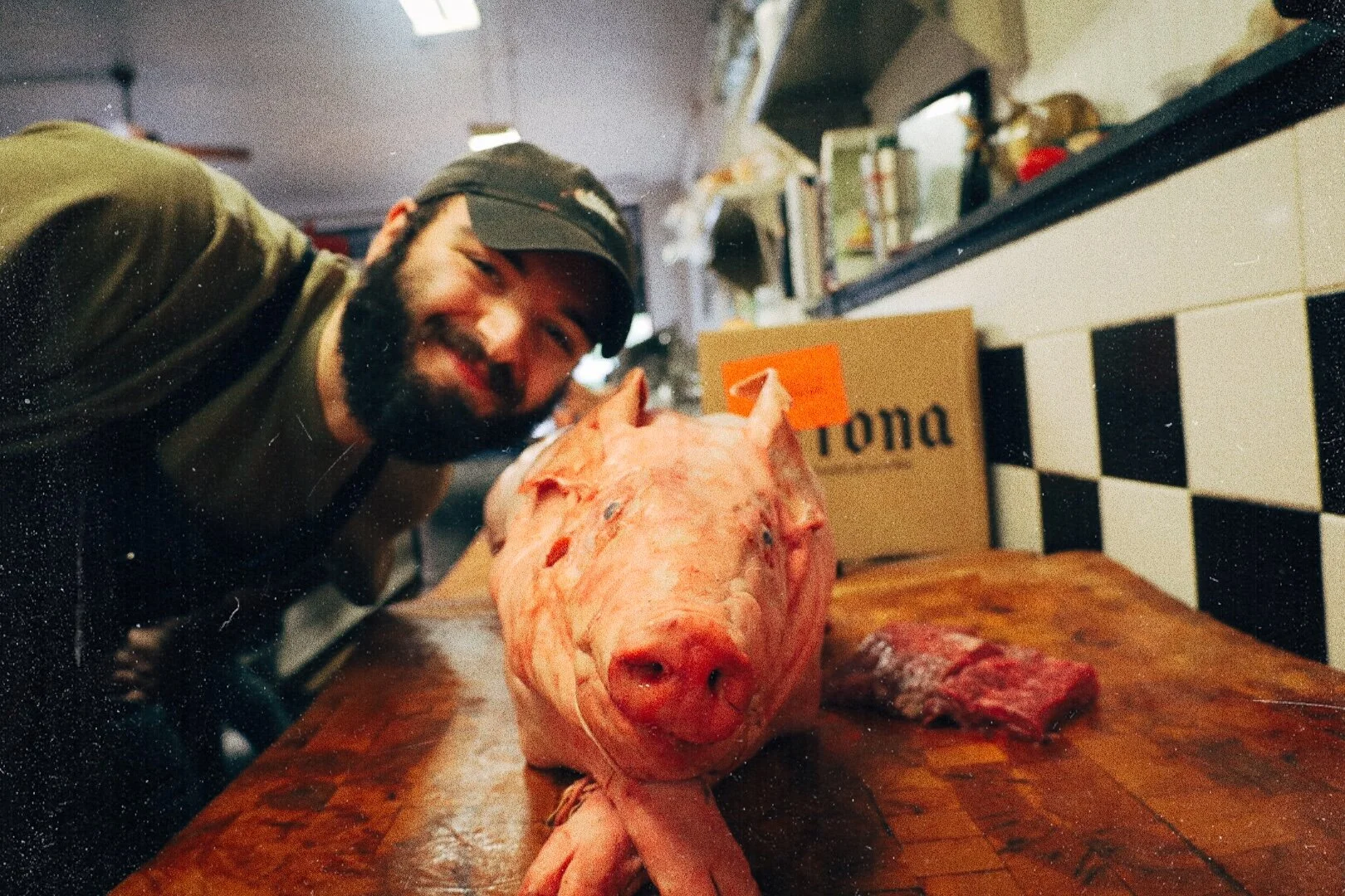 A man with a beard, smiling and leaning towards the camera, is in a butcher shop or meat market next to a pig's head on a wooden cutting board.