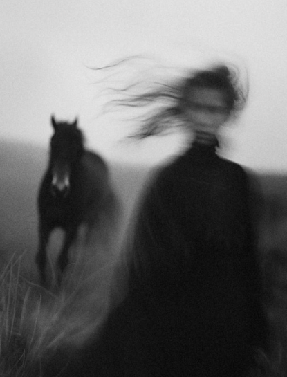 Ein verschwommenes Schwarz-Weiß-Foto einer Person mit langem Haar, die im Wind steht, und einem galoppierenden Pferd im Hintergrund.