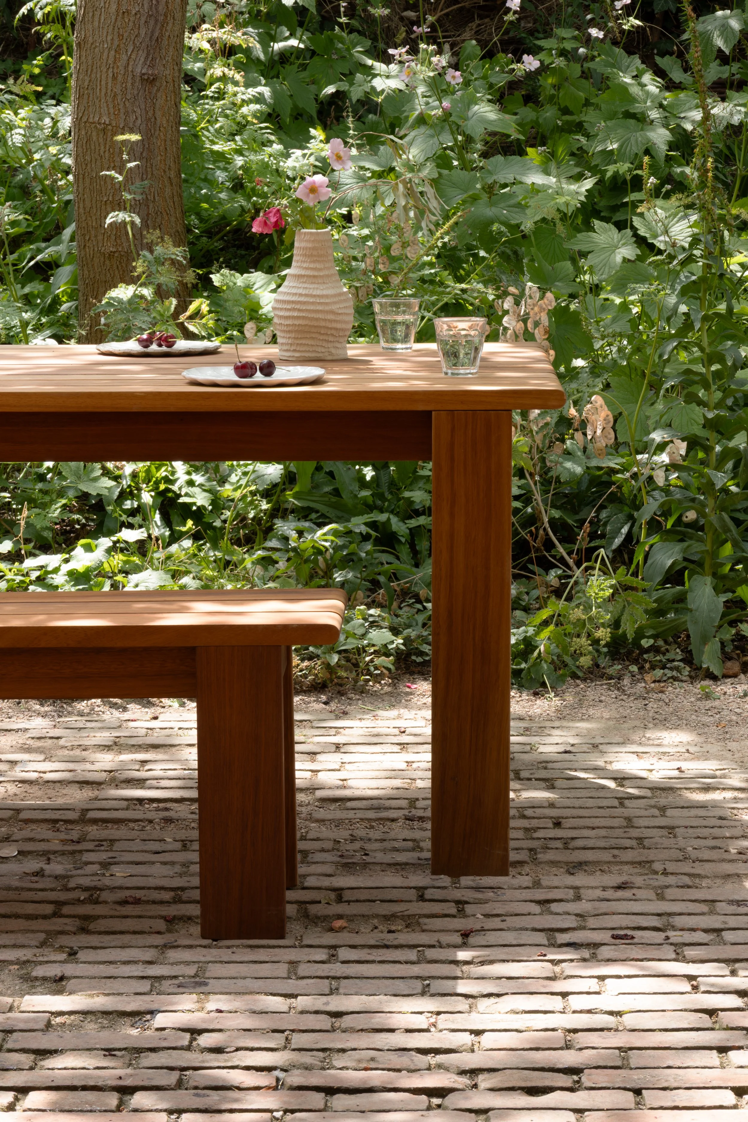 A wooden outdoor table with a ceramic vase holding pink and white flowers, two glasses of water, and two plates of cherries, set on a brick patio surrounded by greenery and trees.