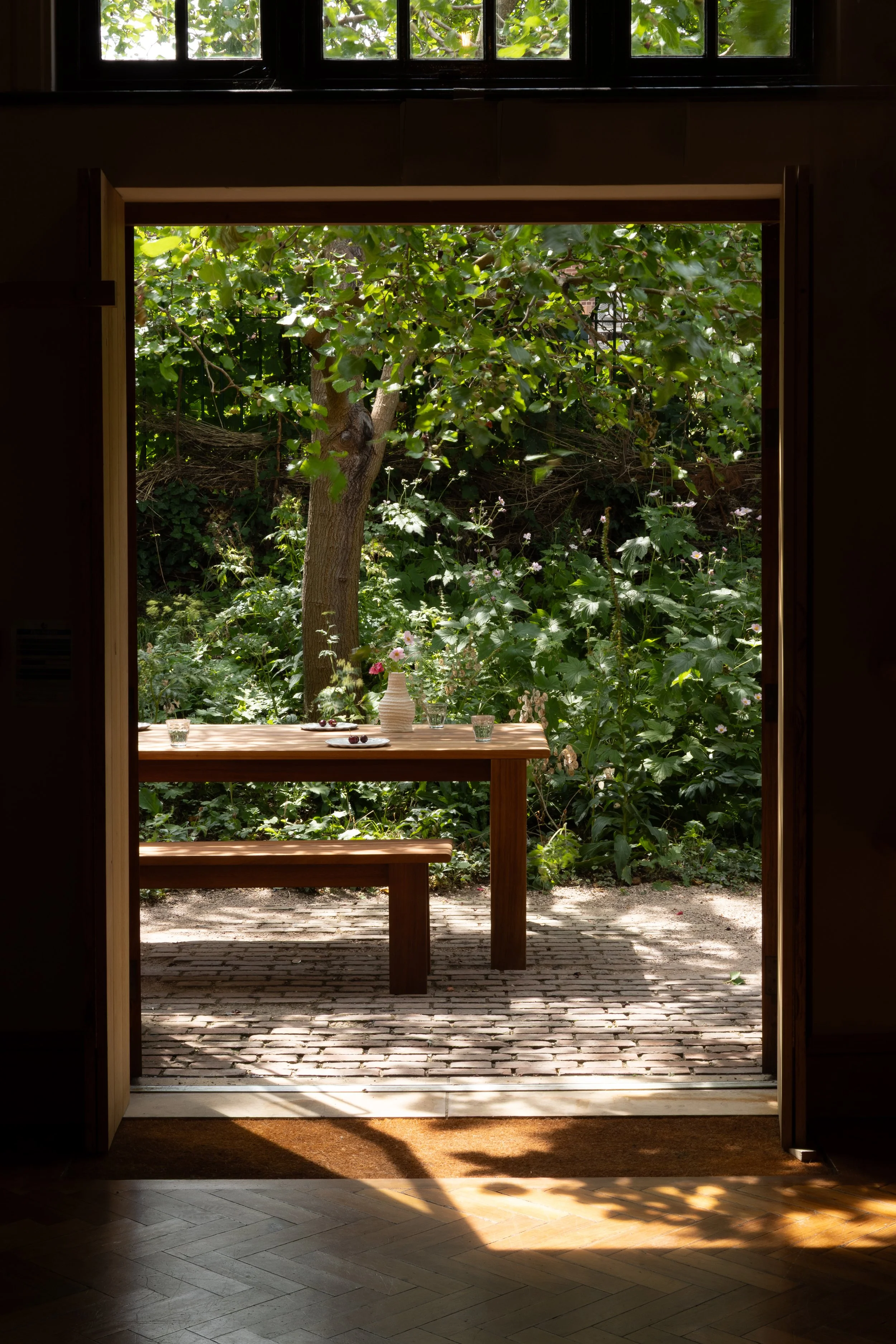 Open door leading to a garden with lush green trees and plants, with a wooden table set with a white vase and small dishes, outdoors on a cobblestone patio.