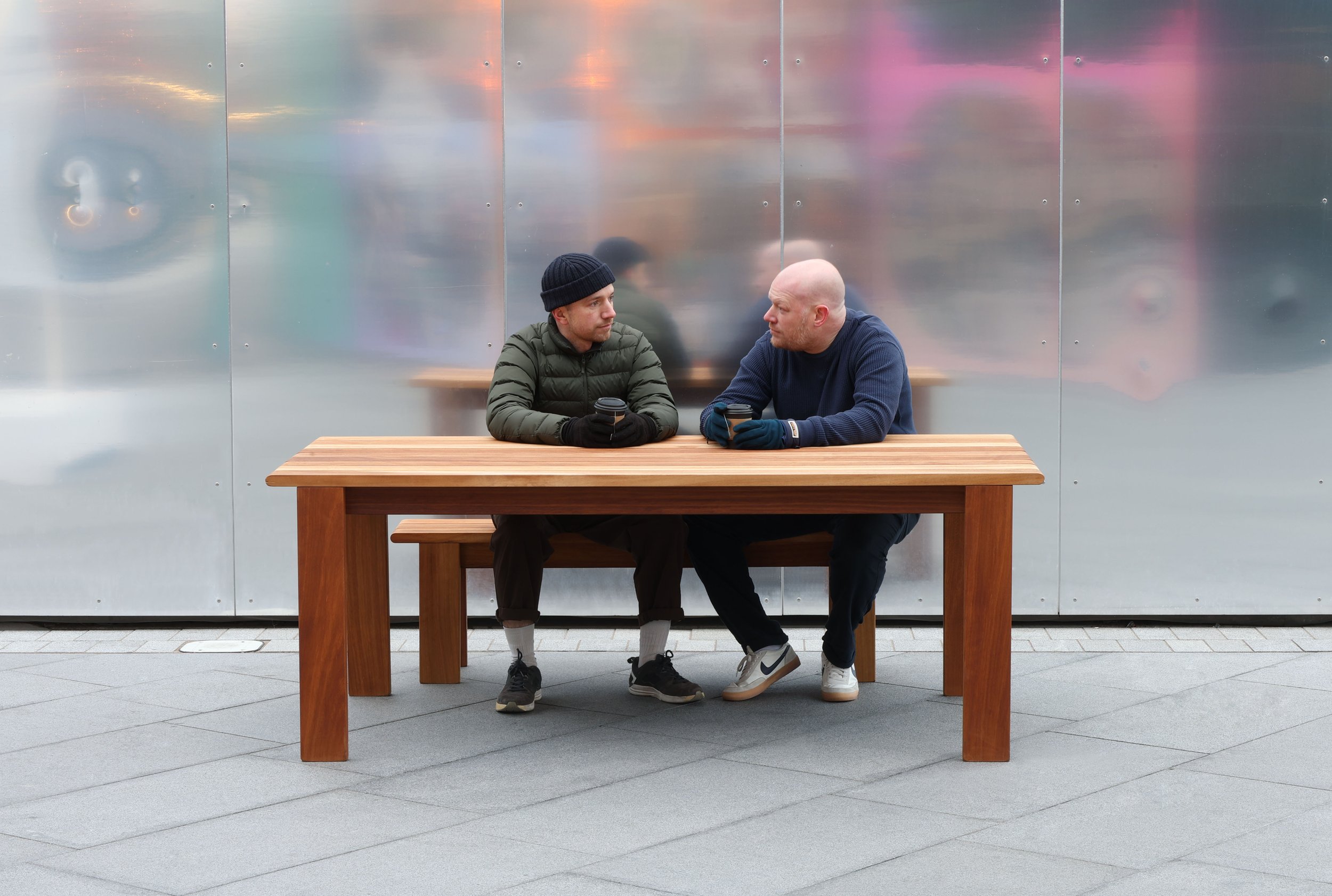 Two men sitting on a wooden bench outdoors, having a conversation while holding coffee cups, in front of a metallic wall.