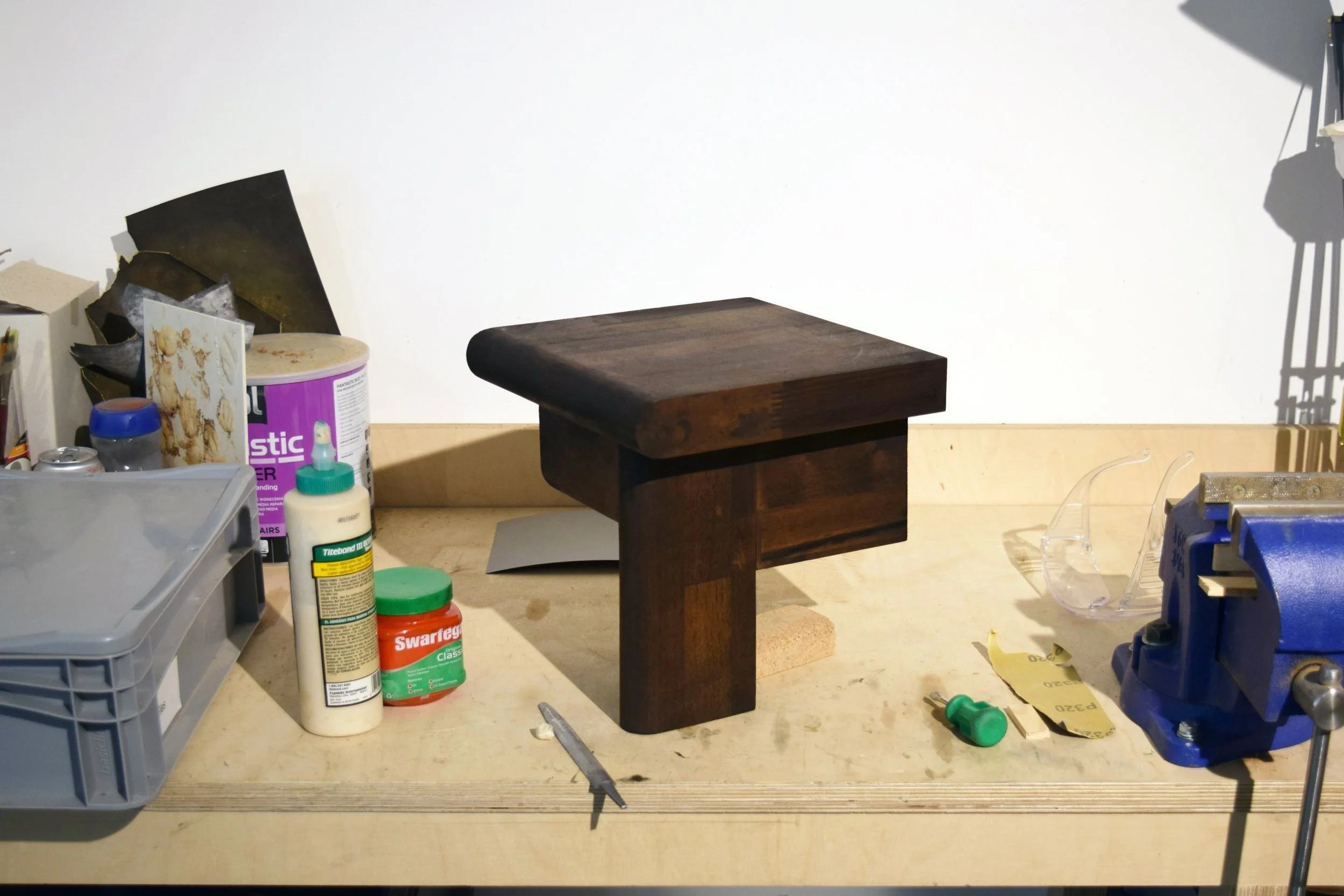 A cluttered table with various objects, including a small dark wooden table, bottles, a glue stick, a needle, and a transparent plastic container, set against a white wall with shadows.