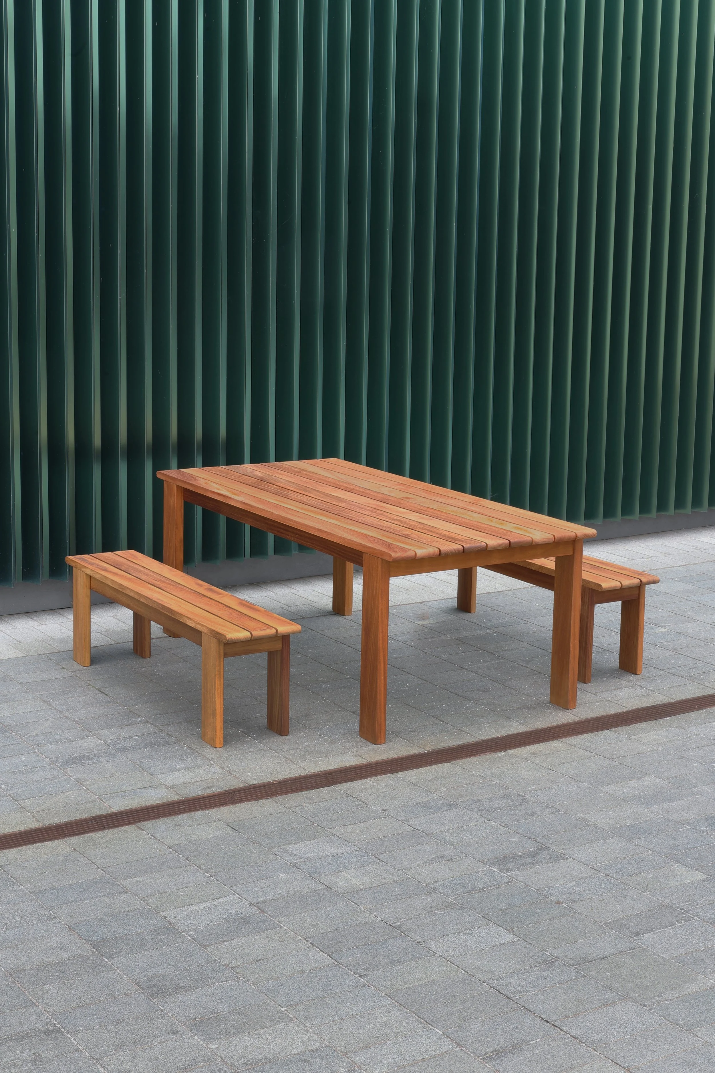 Wooden picnic table with attached benches on a gray tiled outdoor floor. A green corrugated metal wall is in the background.