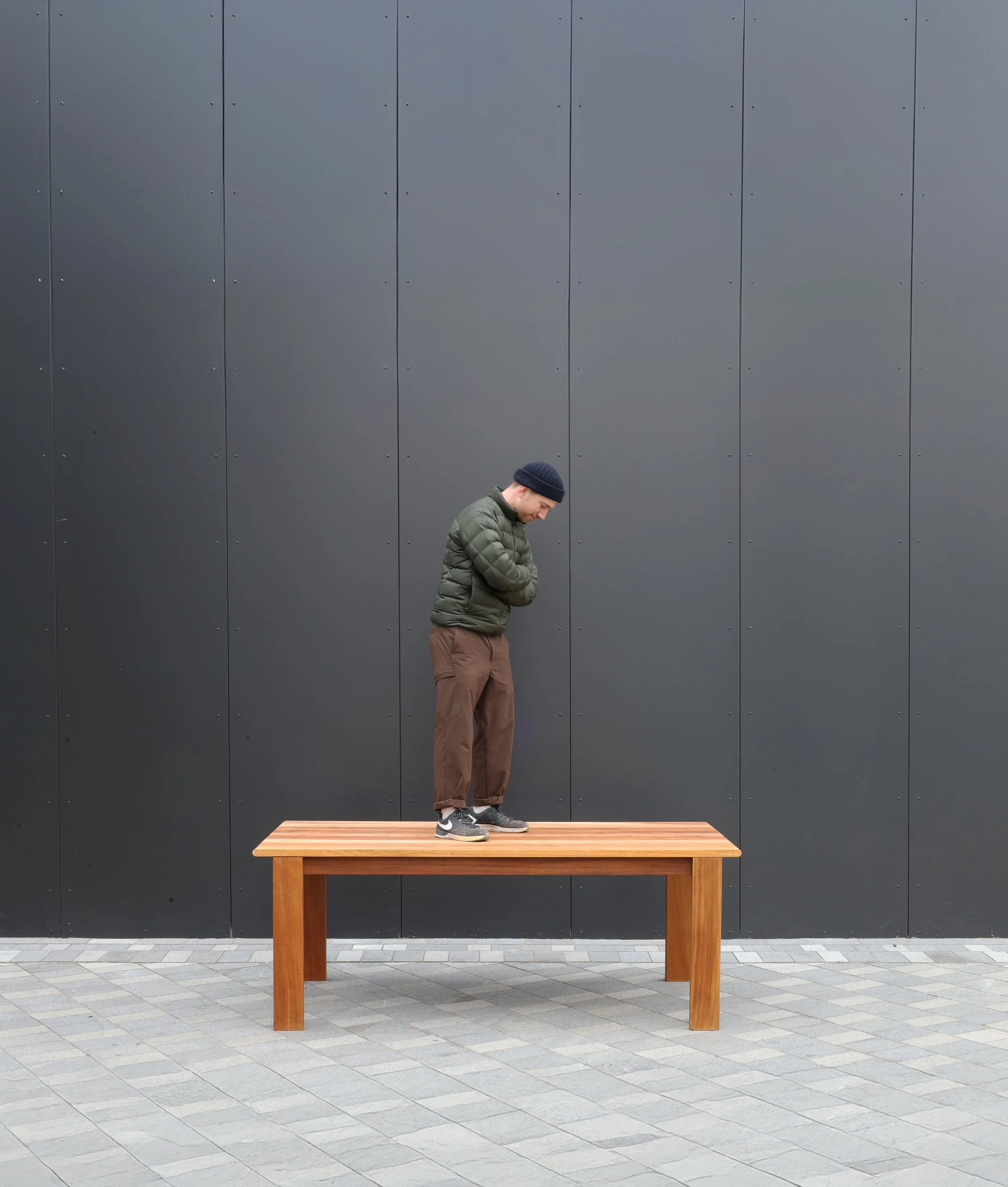 A person with a dark blue beanie, green jacket, and brown pants standing on a wooden table in front of a dark gray wall, with their head bowed down and arms crossed.