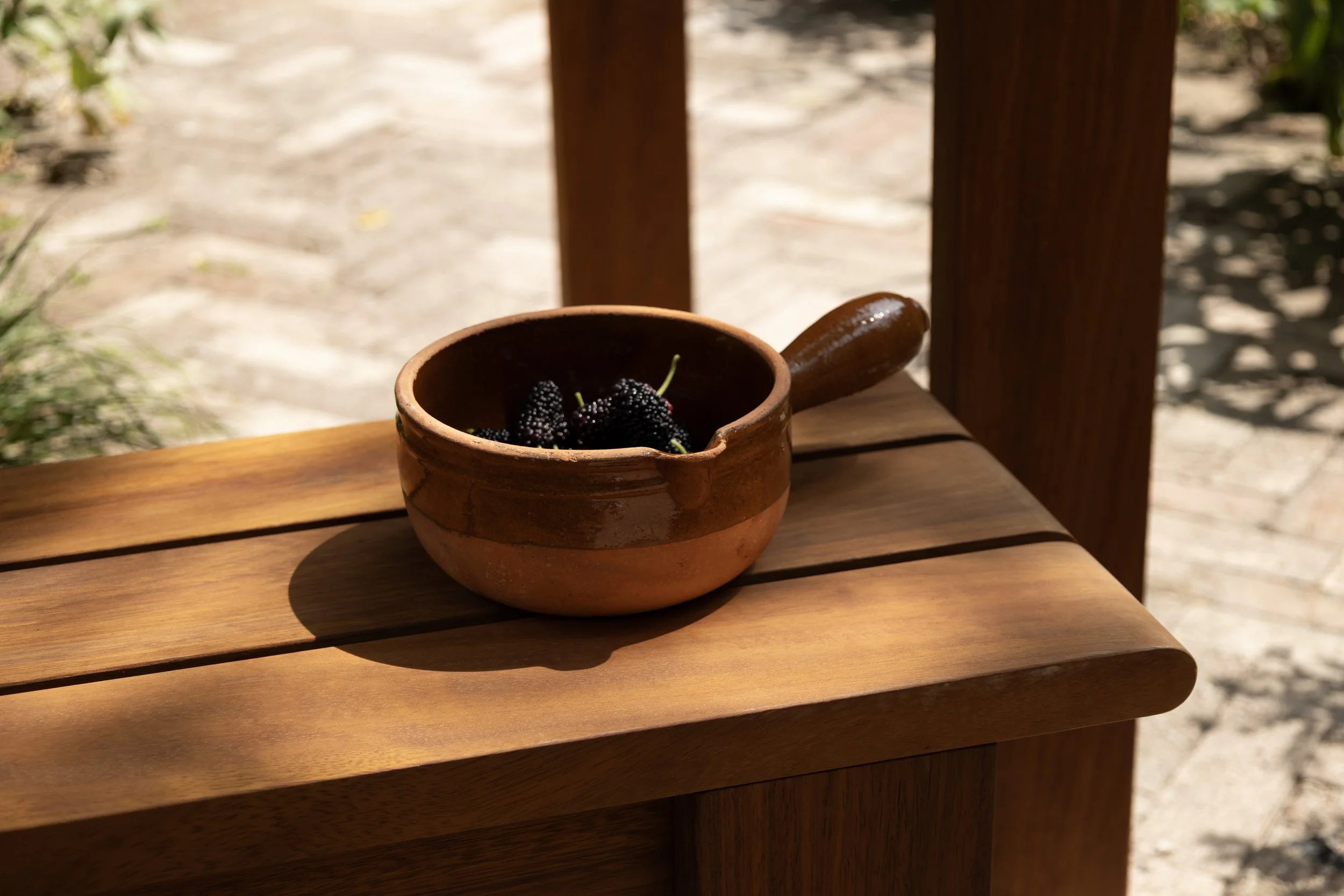 A small clay bowl with blackberries on a wooden table outside.